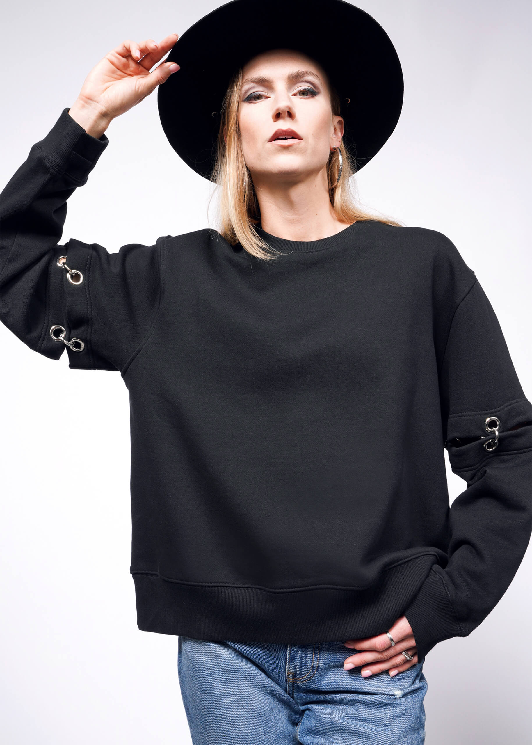 A person in a black wide-brimmed hat, the Debbie Harry X Wildfang Convertible Sweatshirt with metallic loops and detachable sleeves, and blue jeans stands against a white background, raising one hand to adjust the hat.