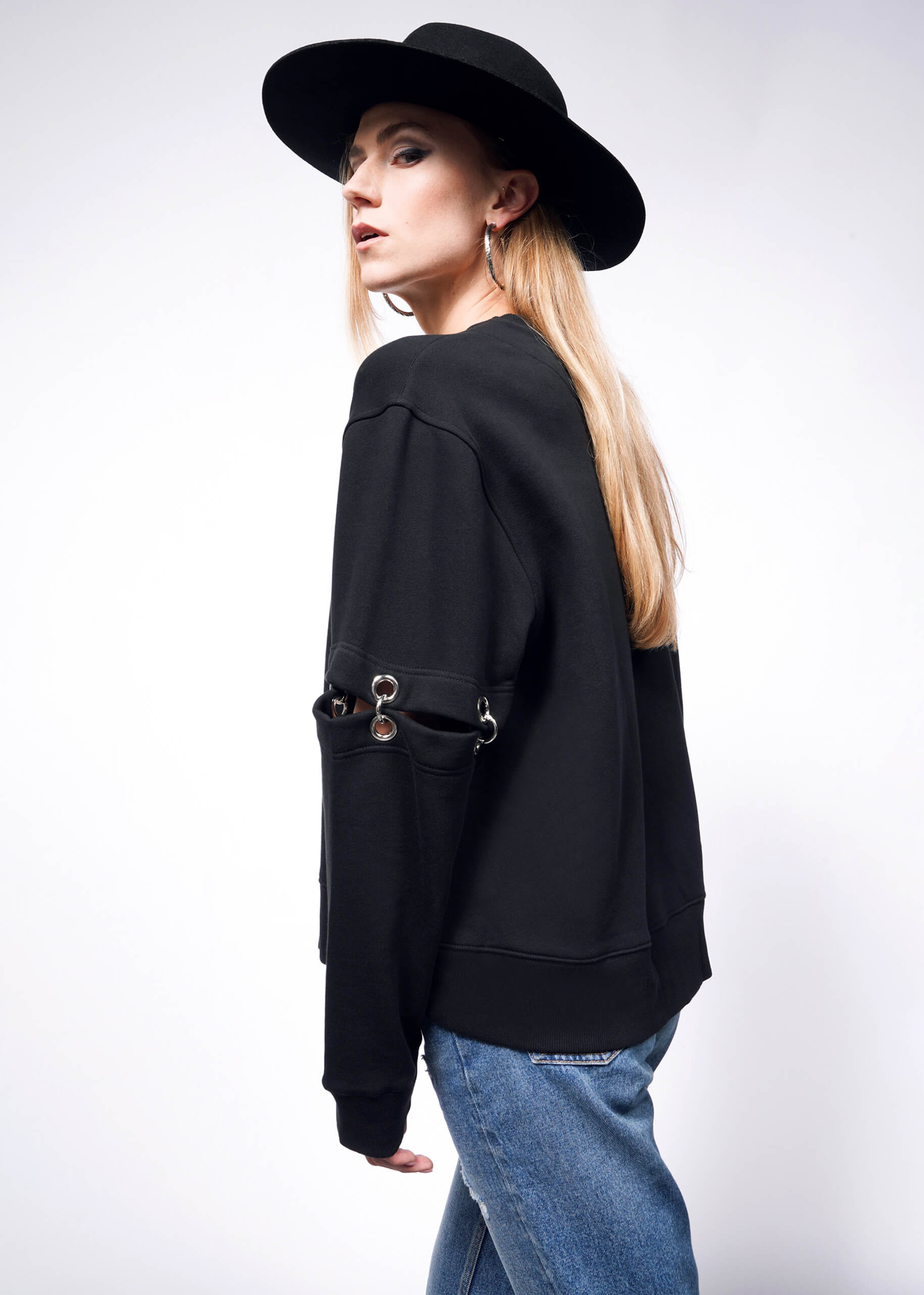 A person with long blonde hair, styled like Debbie Harry X Wildfangs edgy look, wears a black hat and the Convertible Sweatshirt featuring metal ring embellishments and removable sleeves. They pose against a white background in blue jeans, glancing over their shoulder.