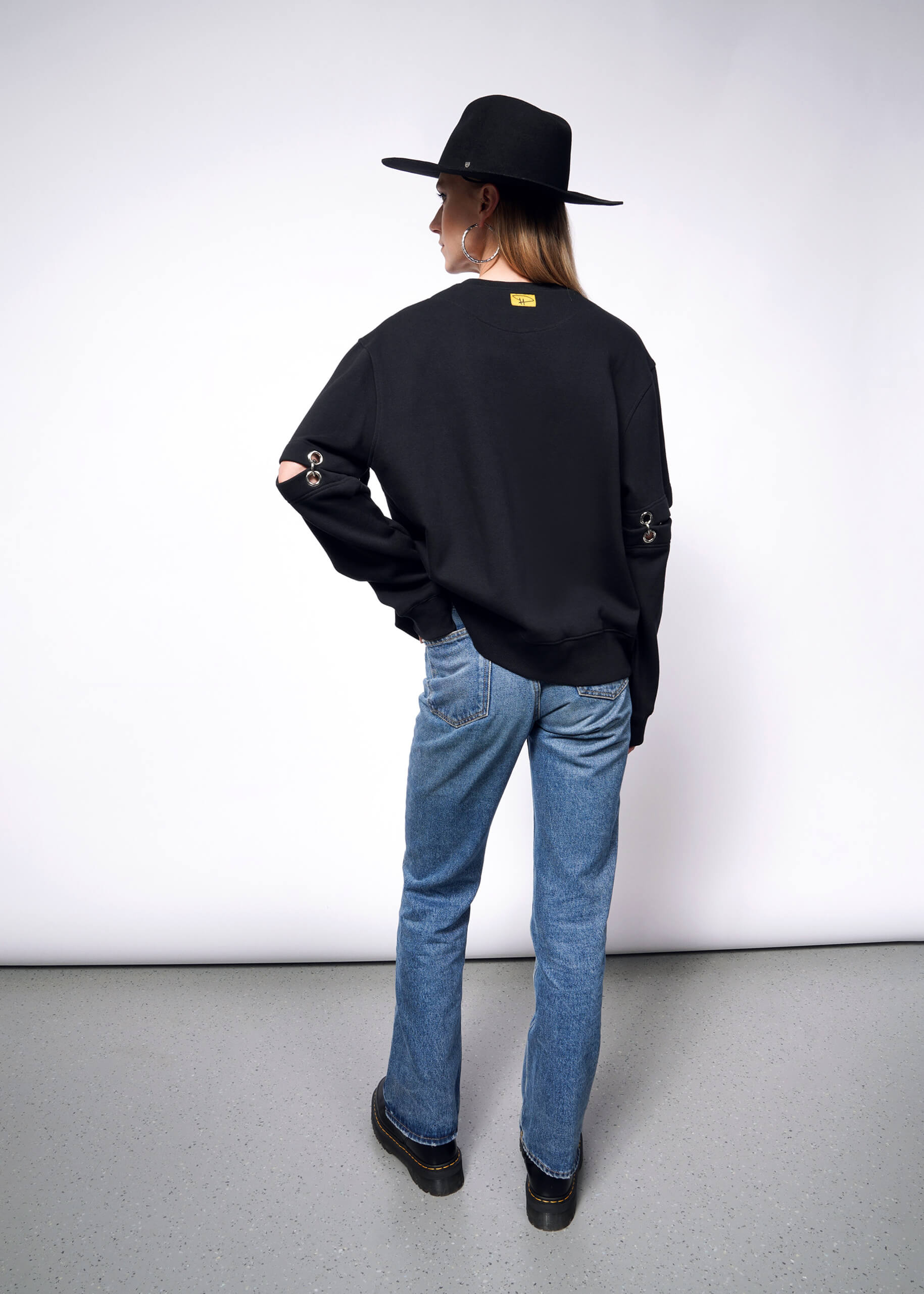 Wearing a Debbie Harry X Wildfang Convertible Sweatshirt with removable sleeves and button details, paired with a wide-brimmed hat and blue jeans, the person stands on a speckled gray floor, facing a plain white wall.