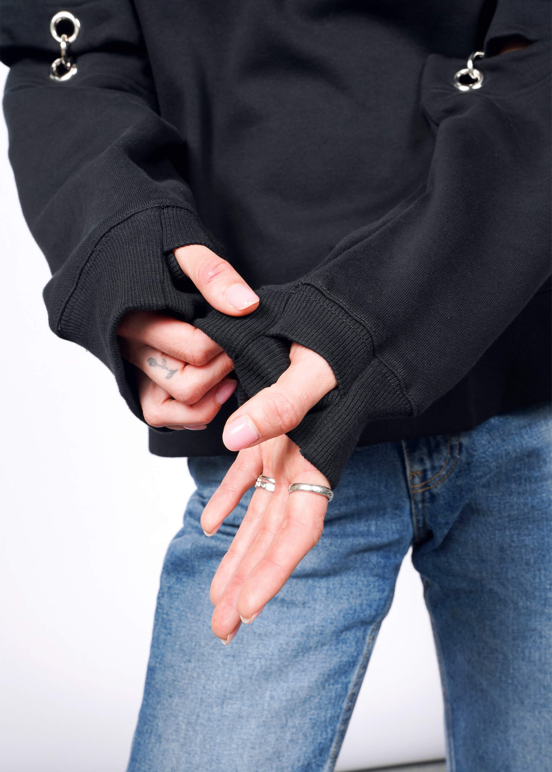 Wearing a black Debbie Harry X Wildfang Convertible Sweatshirt with blue jeans, a person adjusts the ring-adorned removable sleeve, revealing tattooed hands and silver rings against a plain white background.