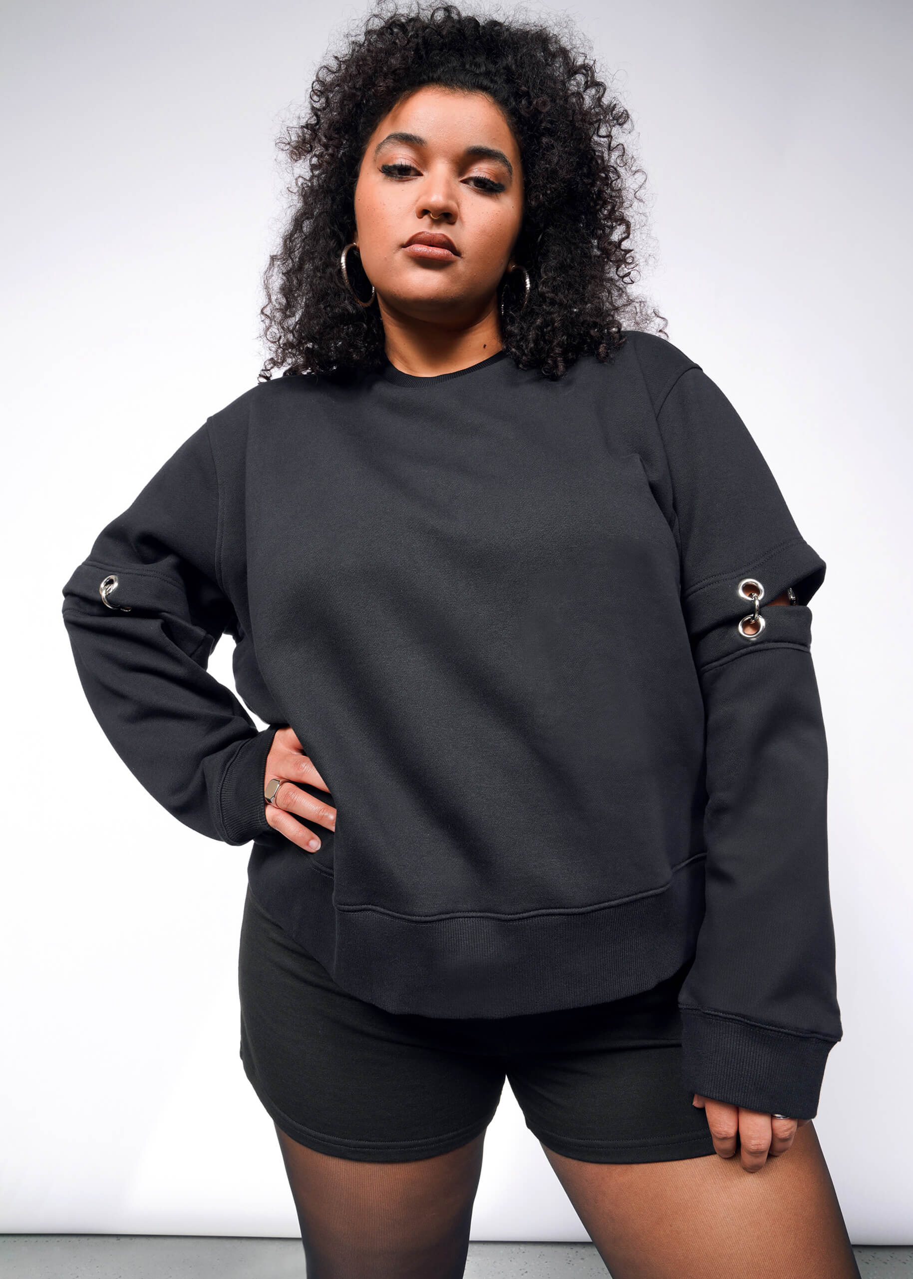 Someone with curly hair poses confidently against a white backdrop in the Debbie Harry X Wildfang Convertible Sweatshirt, showcasing its metal eyelet details and removable sleeves, paired with matching black shorts. They convey a strong, self-assured vibe complemented by hoop earrings.