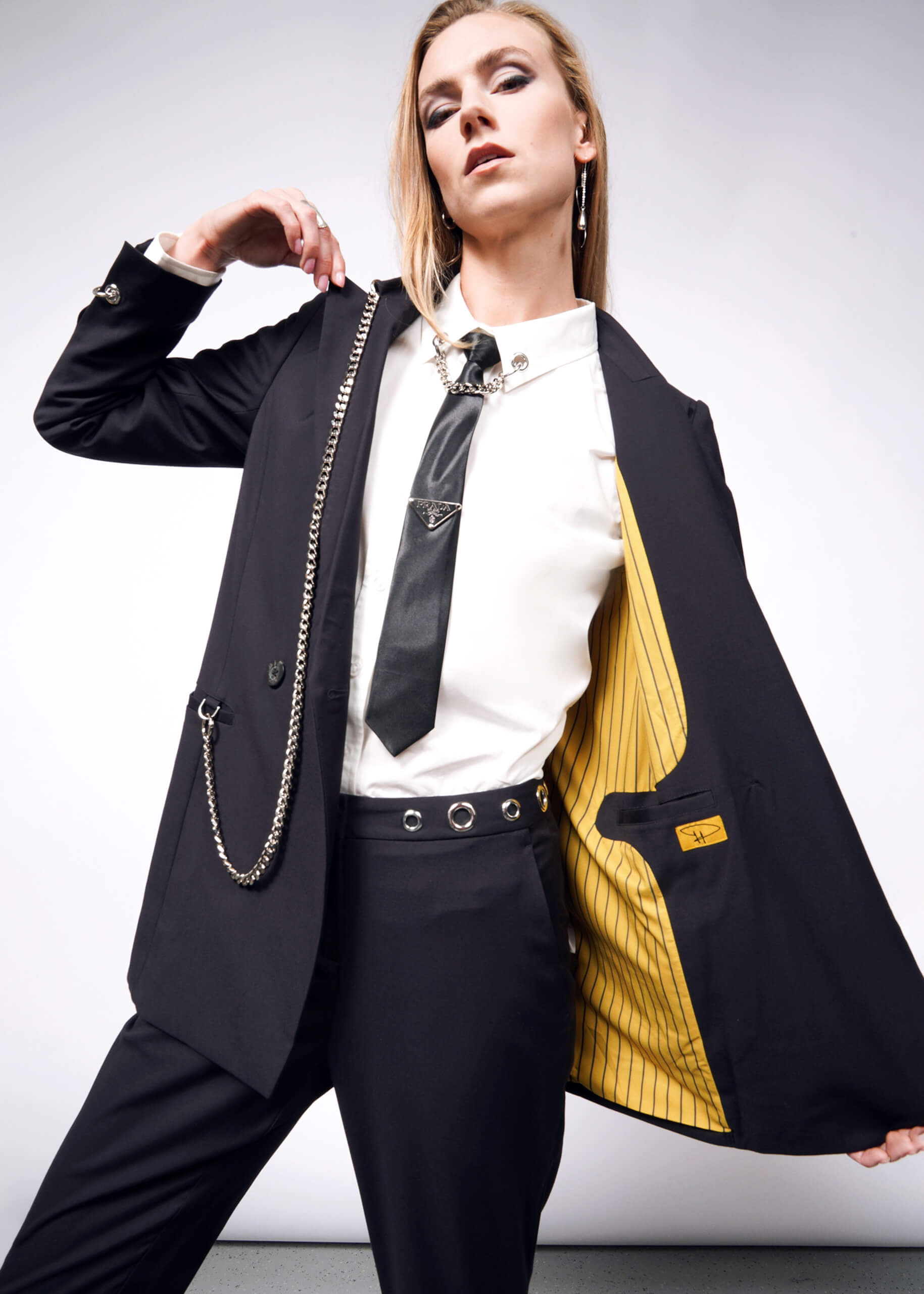 A stylish person poses confidently in a black suit with a white shirt and tie, showcasing the Debbie Harry X Wildfang Double Breasted Blazers striking yellow lining. The outfit is perfectly accessorized with chains and exquisite metal detailing.