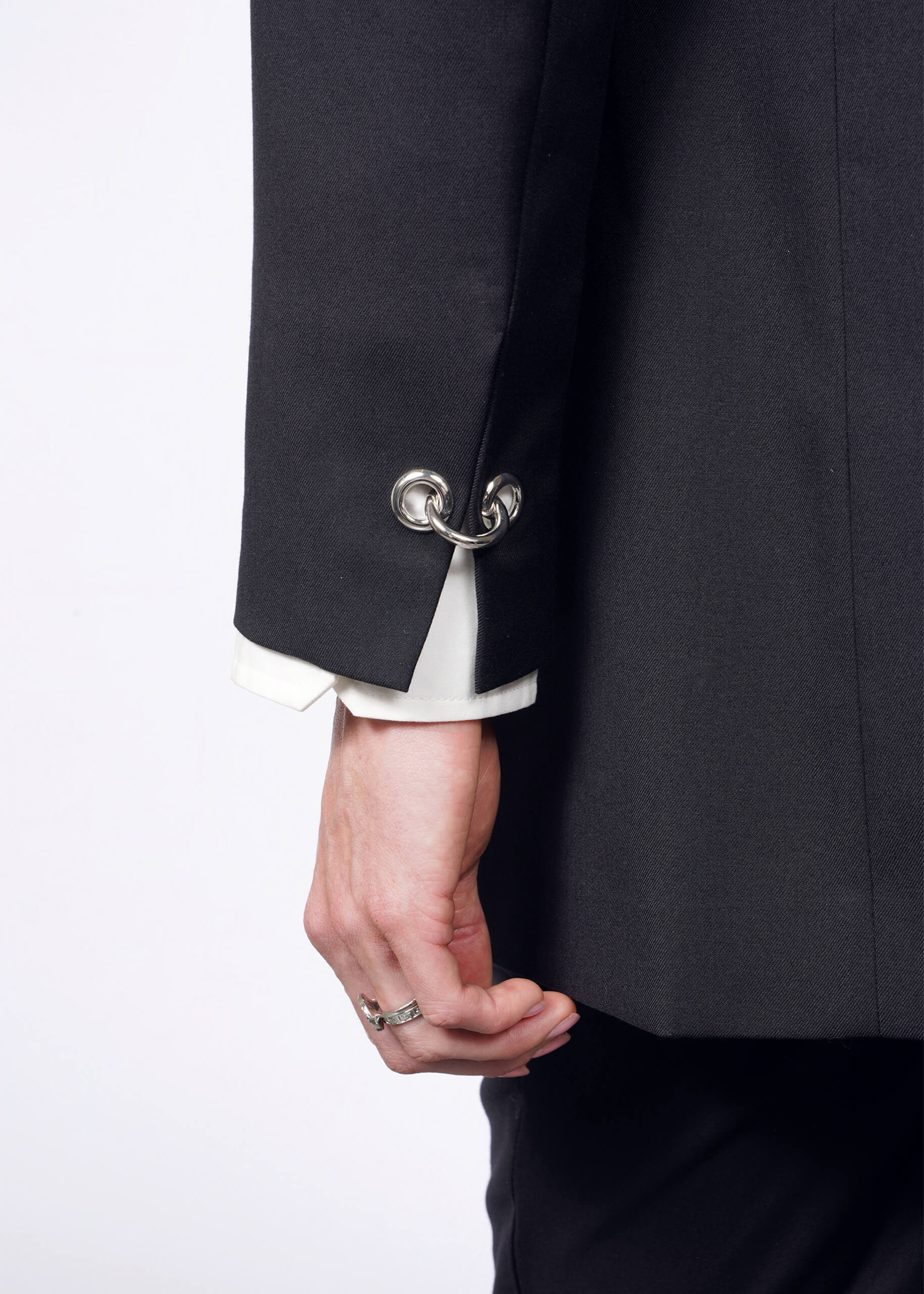 A person wears the sleek black Debbie Harry X Wildfang Double Breasted Blazer featuring metal hoop cufflinks while holding the lapel. A white shirt sleeve shows, paired with a silver ring on their middle finger, contrasting against a white background and highlighting striking metal details.