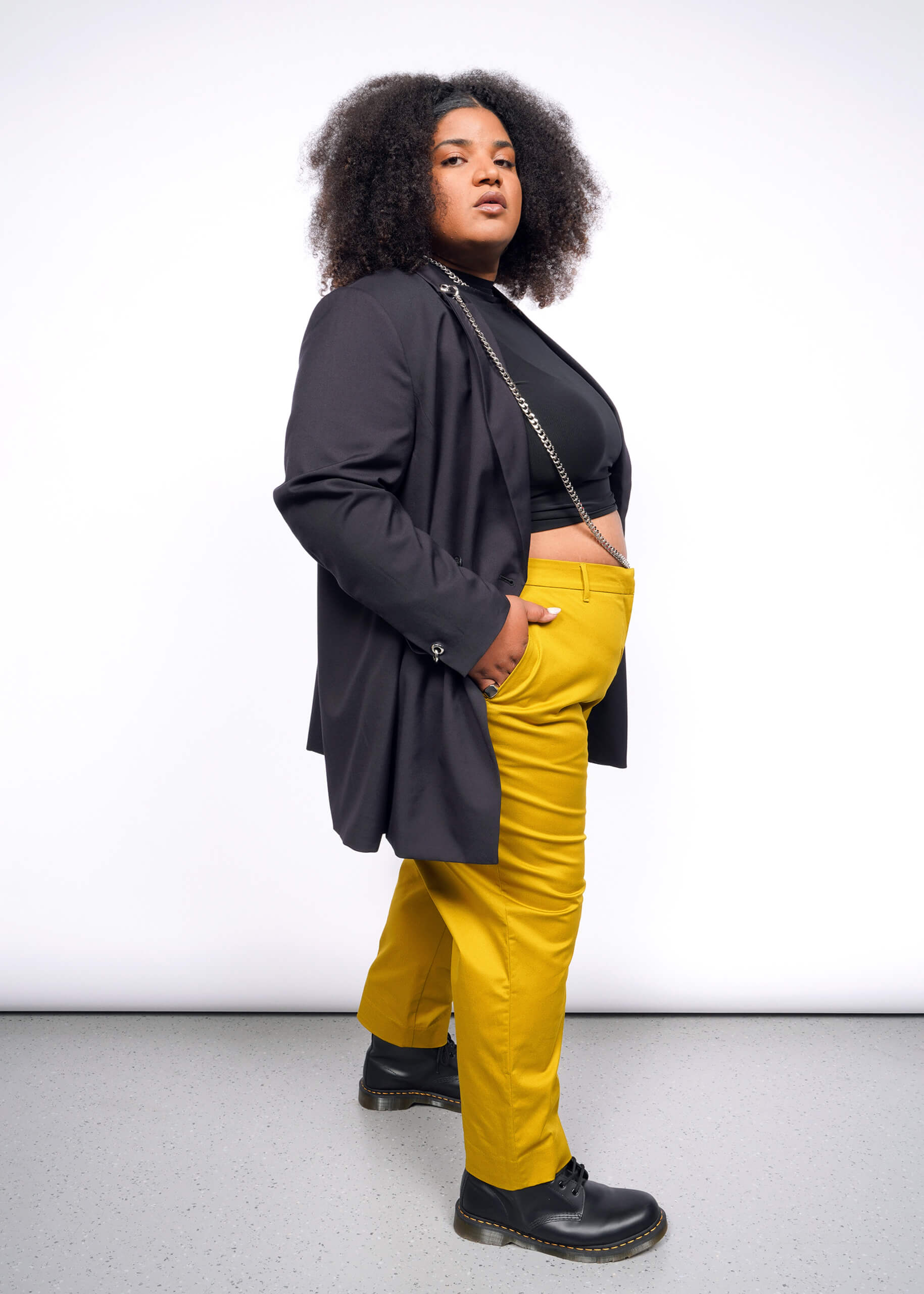 A person with curly hair confidently poses, wearing a Debbie Harry X Wildfang Double Breasted Blazer over a black top and bright yellow pants. The outfit, featuring subtle metal detailing, complements their stance on a grey floor against a plain white backdrop.