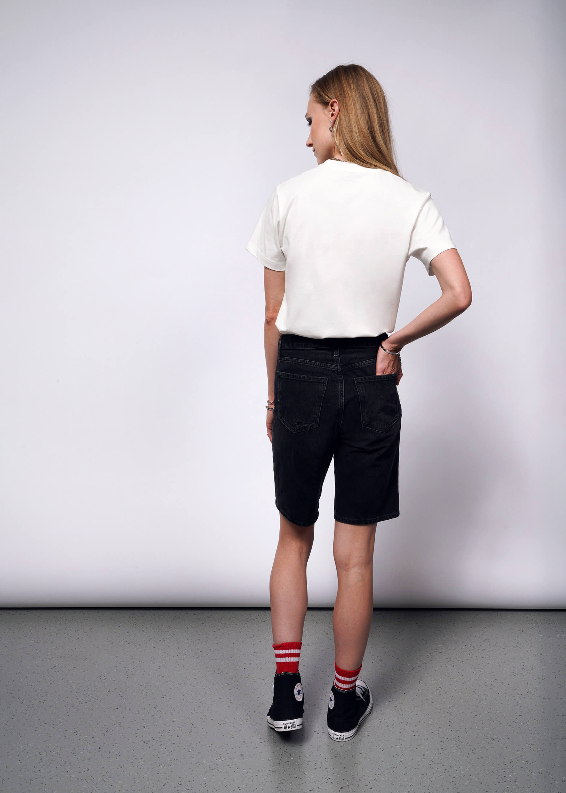 With long hair reminiscent of Debbie Harrys style, a person is seen from the back in a minimalist room with light grey floors and walls. Theyre wearing a Debbie Harry X Wildfang Graphic Tee, black denim shorts, black high-top sneakers, and red striped socks for a punk aesthetic.