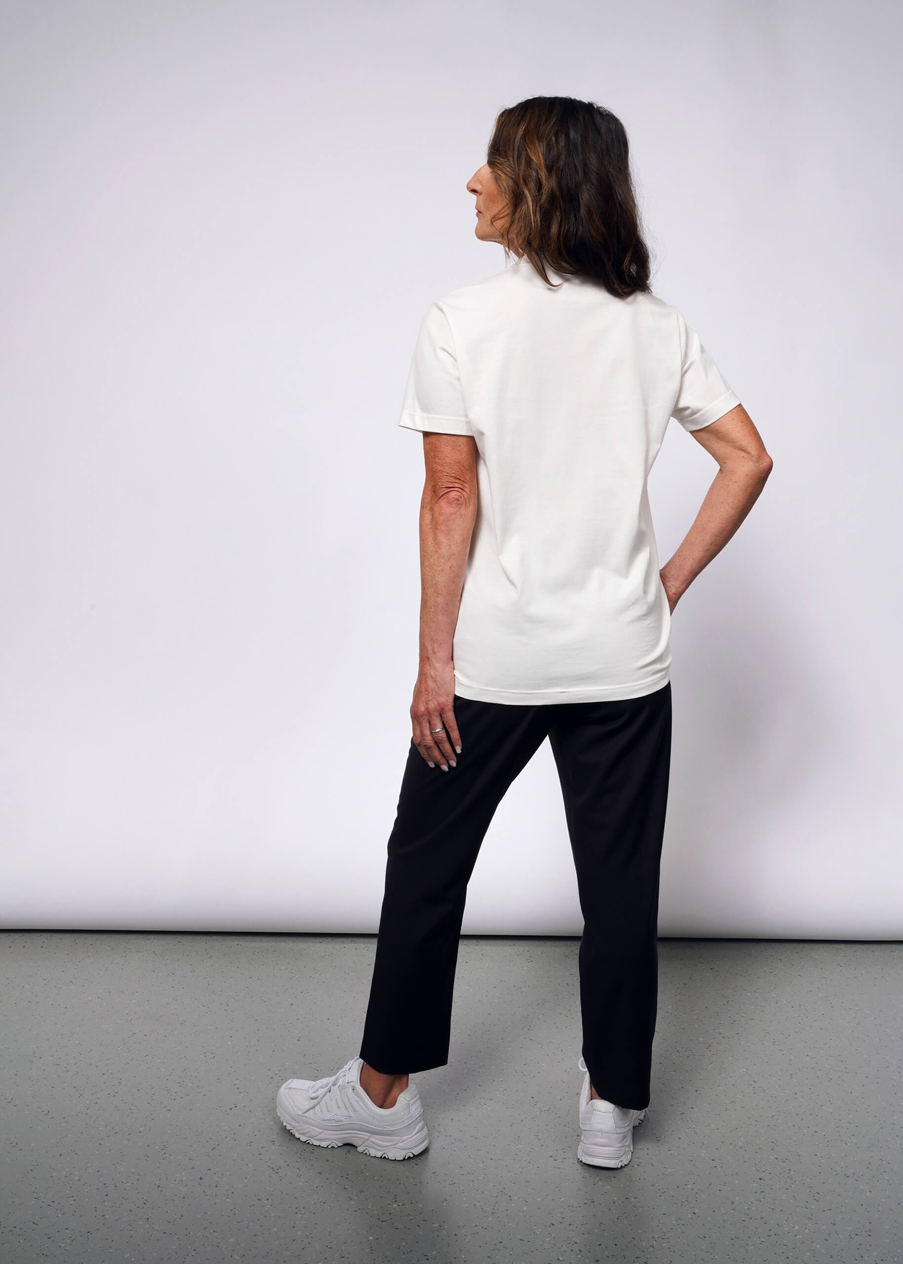 Facing away in a punk style, a woman wears the Debbie Harry X Wildfang Graphic Tee with black pants and white sneakers. Her right hand rests on her hip as she gazes slightly left, creating a modern-day Debbie Harry vibe against the plain light gray background.