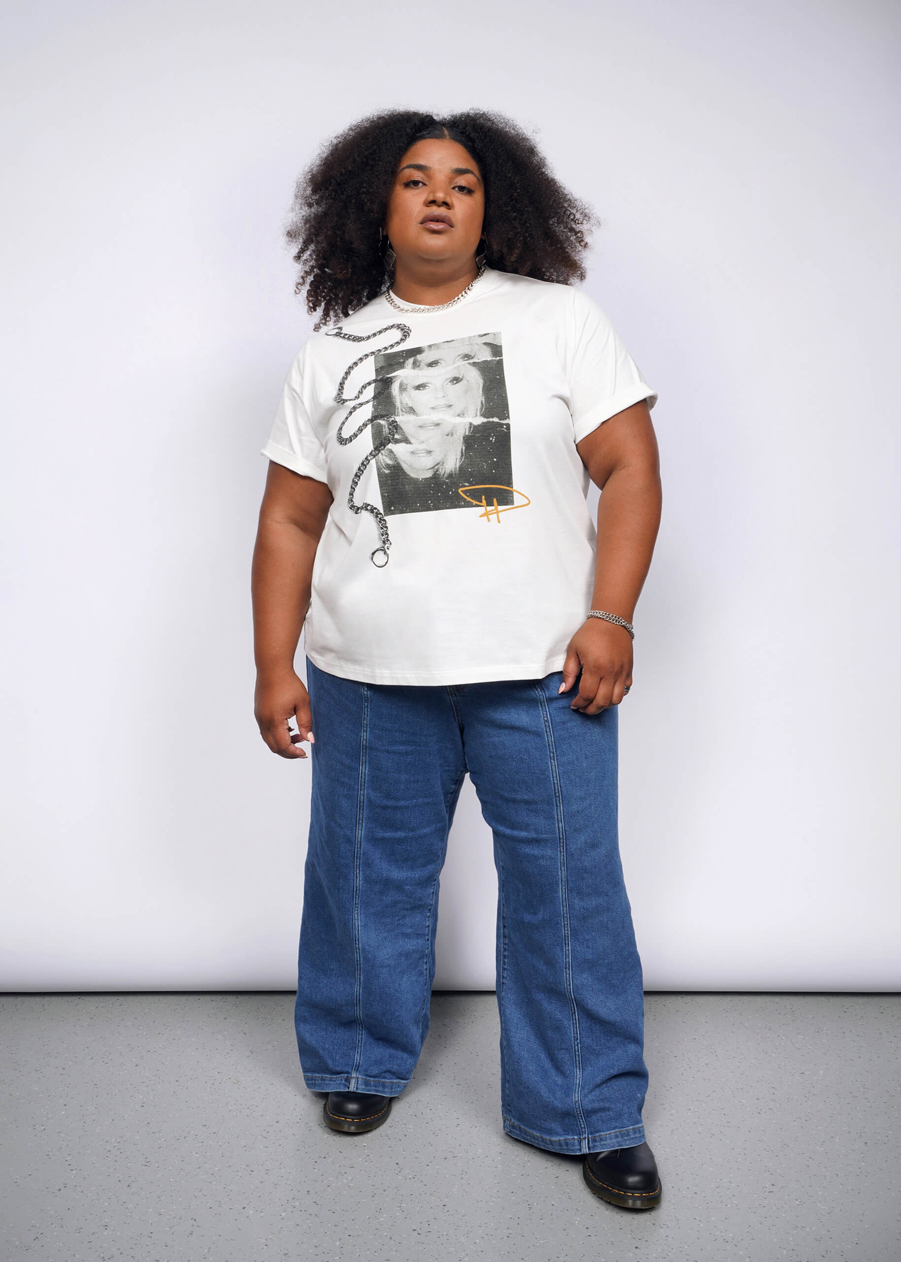 A curly-haired person, embodying a punk aesthetic, stands confidently against a plain background. Theyre wearing the Debbie Harry X Wildfang Graphic Tee with blue jeans and black shoes, exuding the charisma of Debbie Harry.