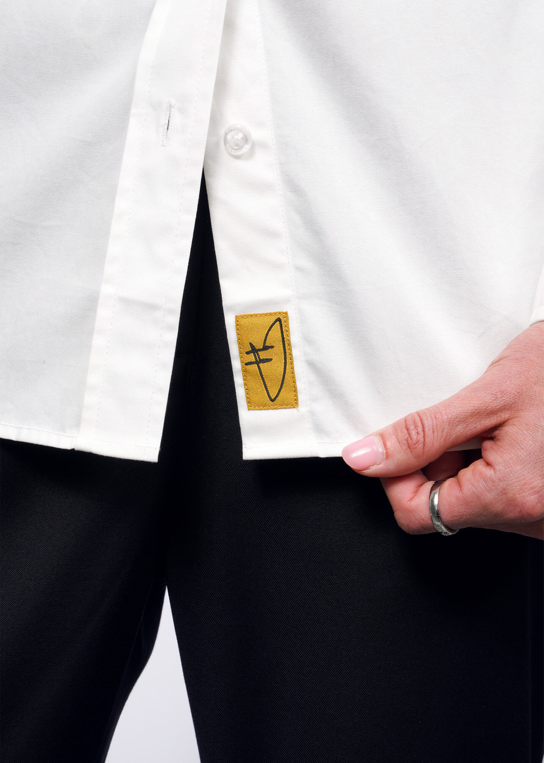 Close-up of a person wearing the Debbie Harry X Wildfang Long Sleeve Button Up and black pants, exuding a punk aesthetic. They hold a yellow tag with an abstract symbol on the shirt. A ring adorns their finger, making a bold statement against the plain background.