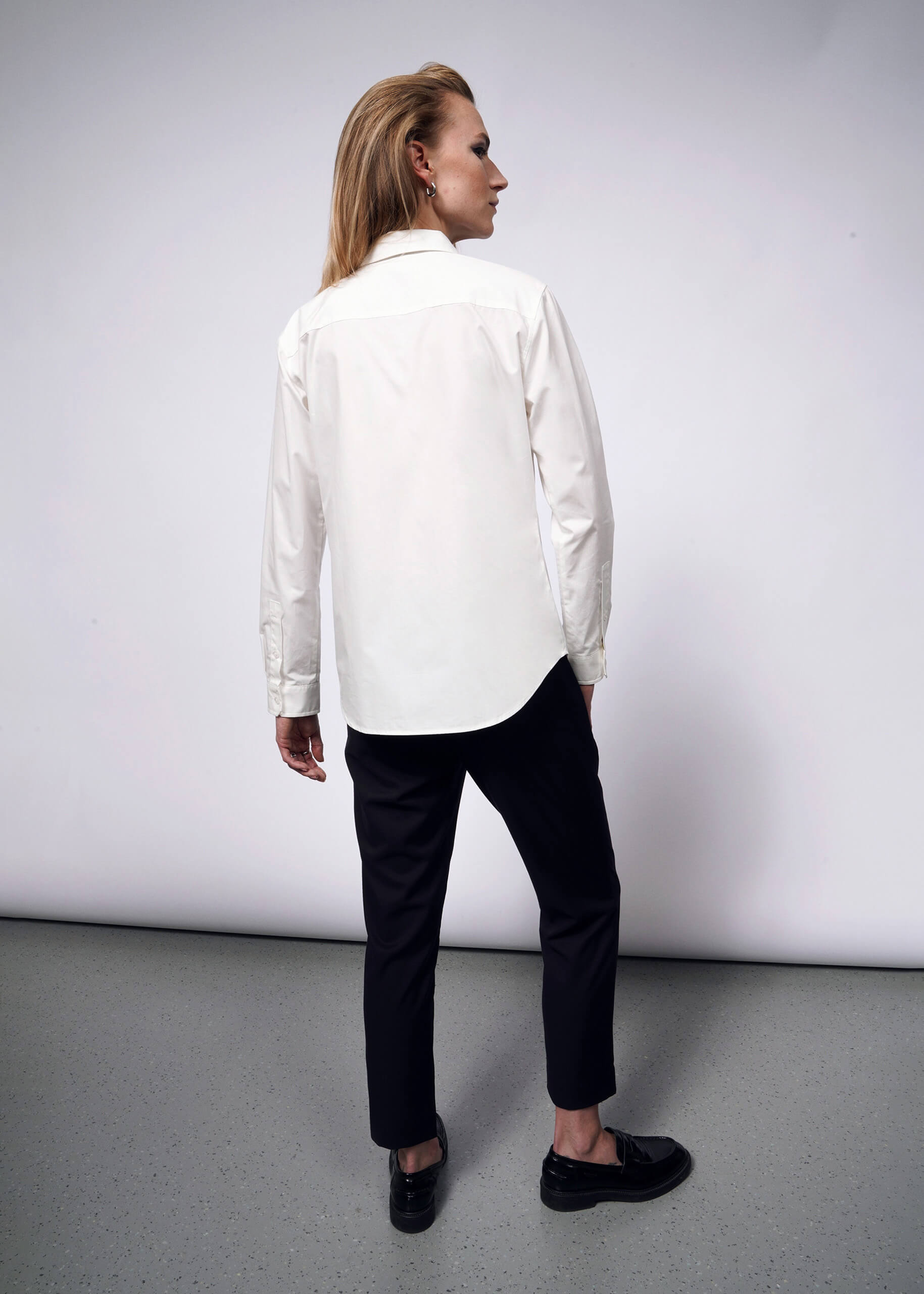 Facing away against a white backdrop, a person with long hair embodies a punk aesthetic in dark pants and the Debbie Harry X Wildfang Long Sleeve Button Up. Their subtle rebellious flair shines as their head turns slightly to the side, while their hands rest relaxed by their sides.