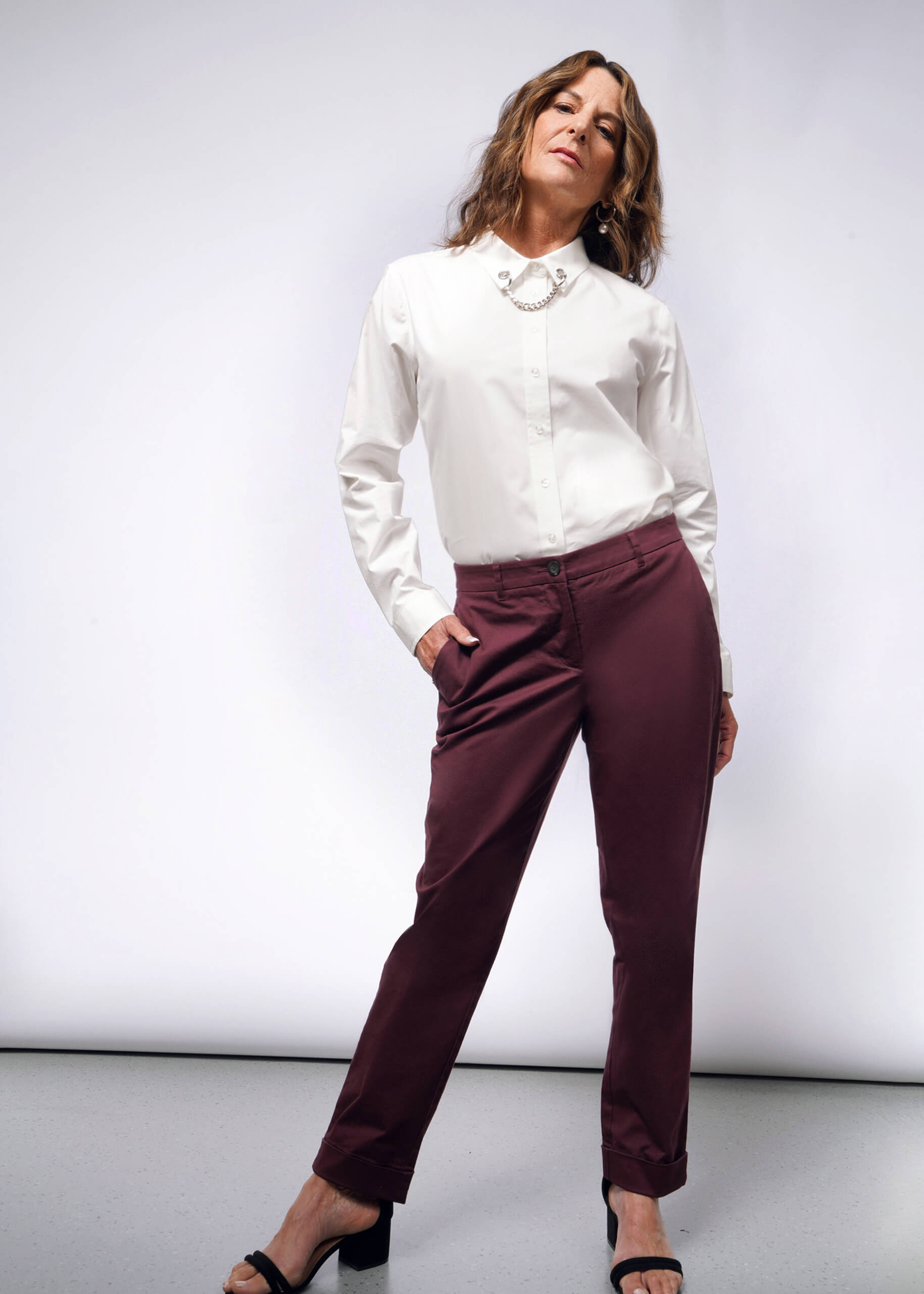 Confidently posing in the Debbie Harry X Wildfang Long Sleeve Button Up, a person pairs it with burgundy pants and black sandals. Standing against a plain backdrop, hands in pockets, they channel a punk aesthetic with a slight smile directed at the camera.