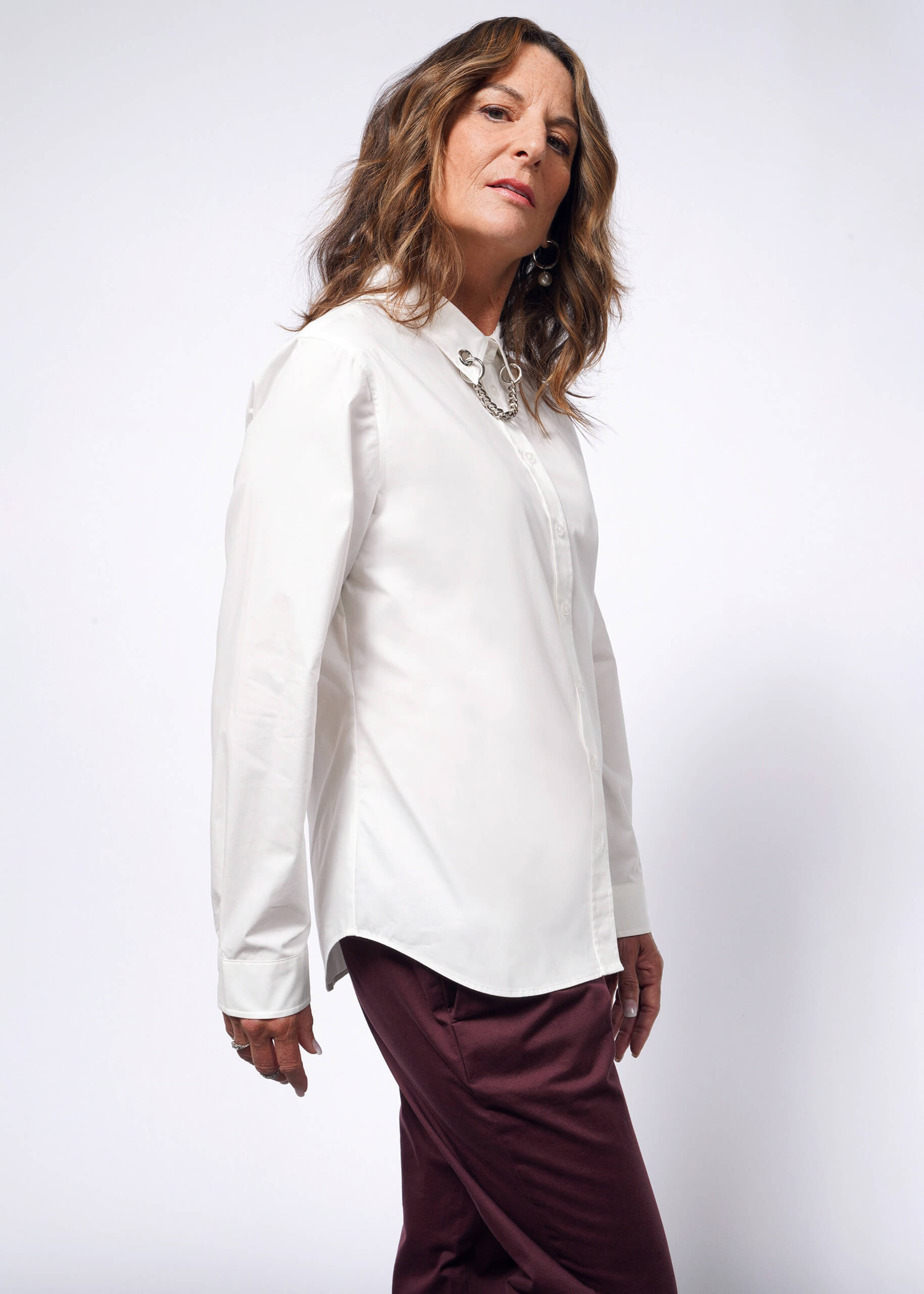 A person with shoulder-length brown hair in a white Debbie Harry X Wildfang Long Sleeve Button Up, featuring a removable chain, and maroon pants stands confidently against a plain background, embodying a subtle punk aesthetic.