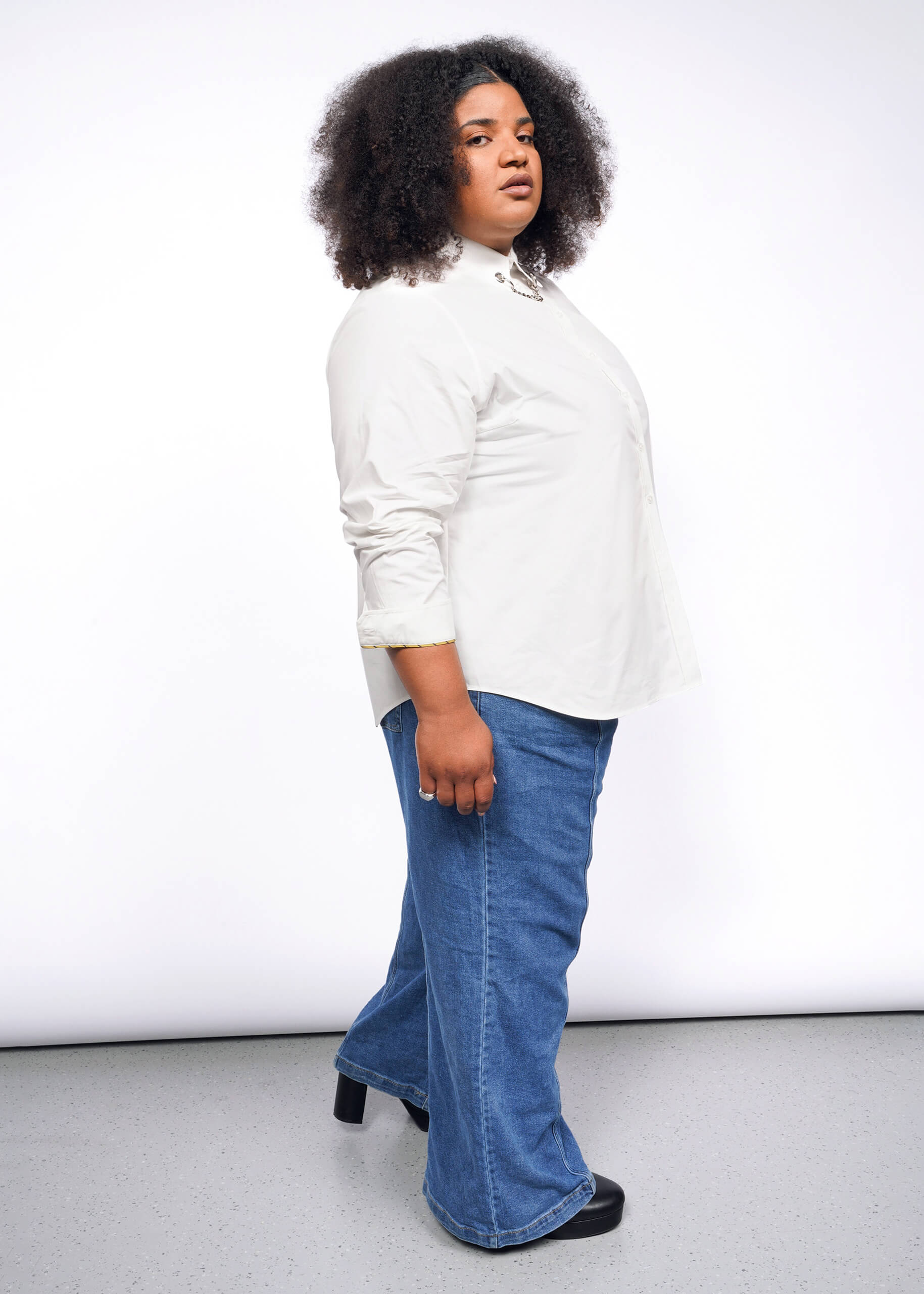 A person with curly hair stands in profile against a white background, channeling a punk vibe. They wear the Debbie Harry X Wildfang Long Sleeve Button Up with removable chain detail, paired with blue jeans and black shoes.