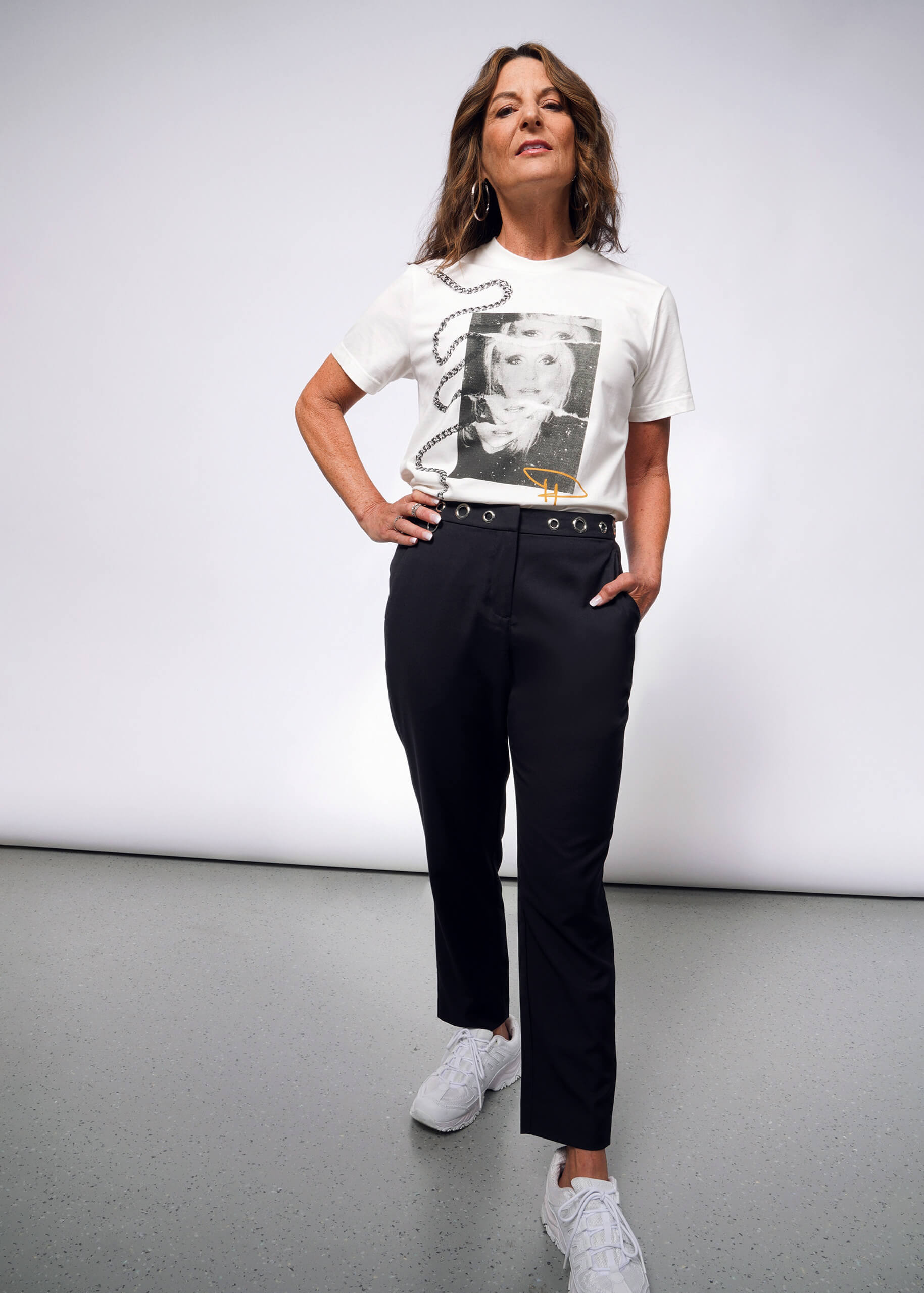 In a bright studio, a woman exudes punk vibes reminiscent of Debbie Harry. She confidently wears the Debbie Harry X Wildfang Slim Crop Pants with a white graphic tee and hoop earrings. Her long brown hair and white sneakers complete her striking look.