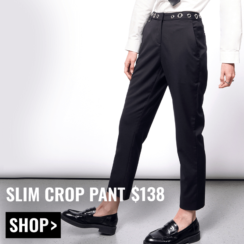 Model pictured from the waist down wearing the black Debbie Harry x Wildfang slim crop pant and black loafers. Text overlay reads SLIM CROP PANT $138 and button underneath reads SHOP. White background.