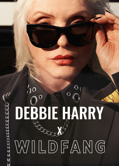 Close up of Debbie Harry looking at camera wearing sunglasses and pieces from the Debbie Harry x Wildfang collection. Text overlay says DEBBIE HARRY X WILDFANG.