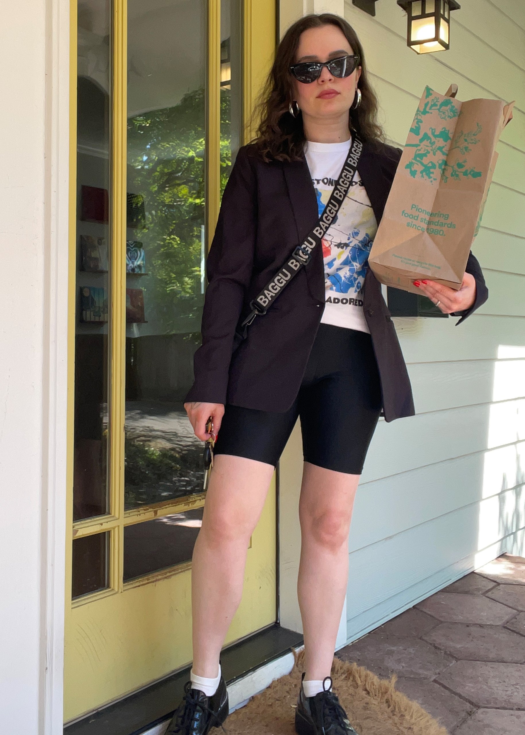 Wearing The Empower Tux Blazer with deep pockets and sunglasses, a woman stands at a doorway holding a paper grocery bag. She pairs her look with a graphic t-shirt, black shorts, shoes, and has a strap bag slung across her shoulder.