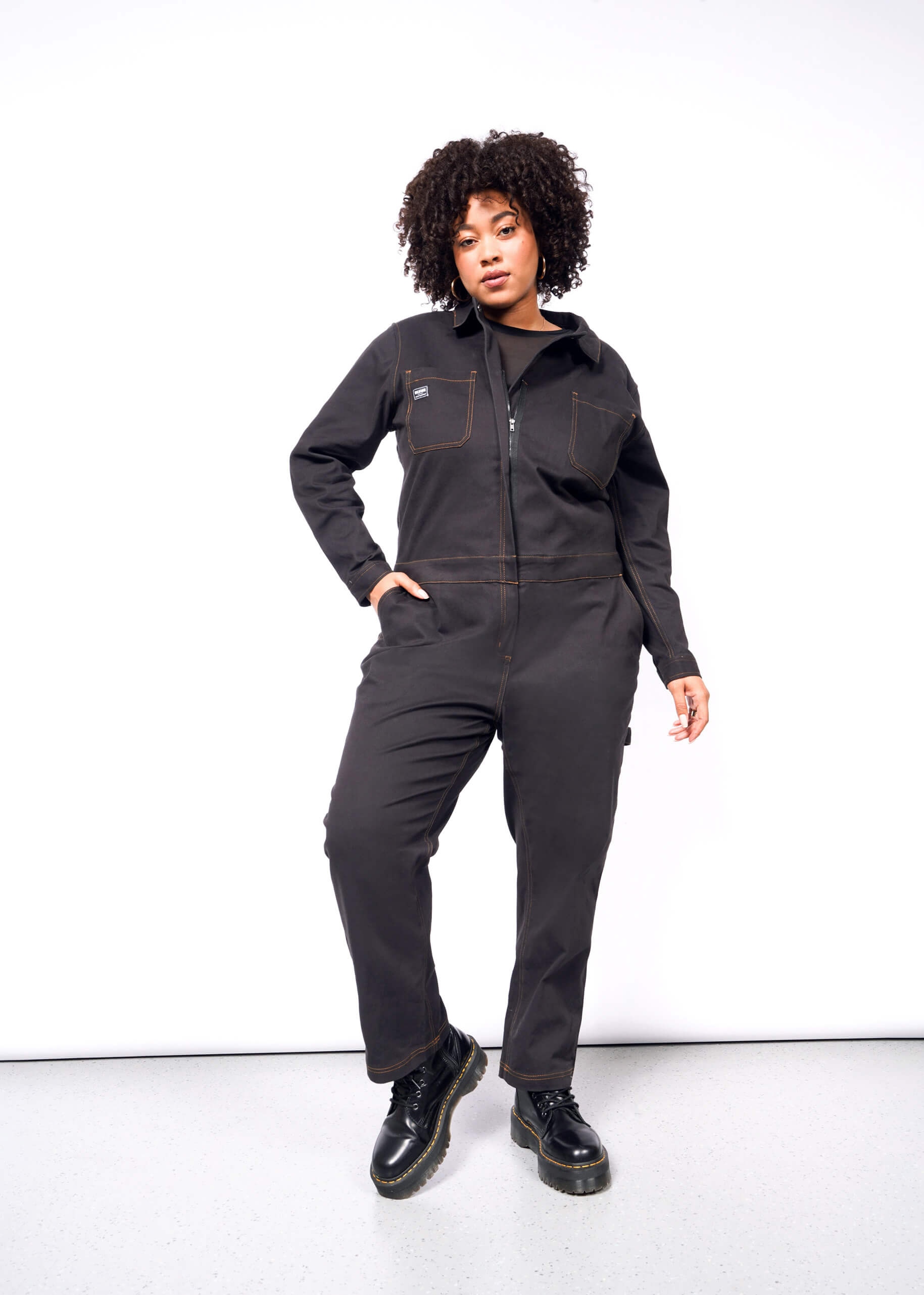 A person with curly hair confidently stands in The Essential Long Sleeve Coverall, made of brushed stretch cotton, while wearing black boots. One hand in the pocket and the other on their hip against a plain white background.