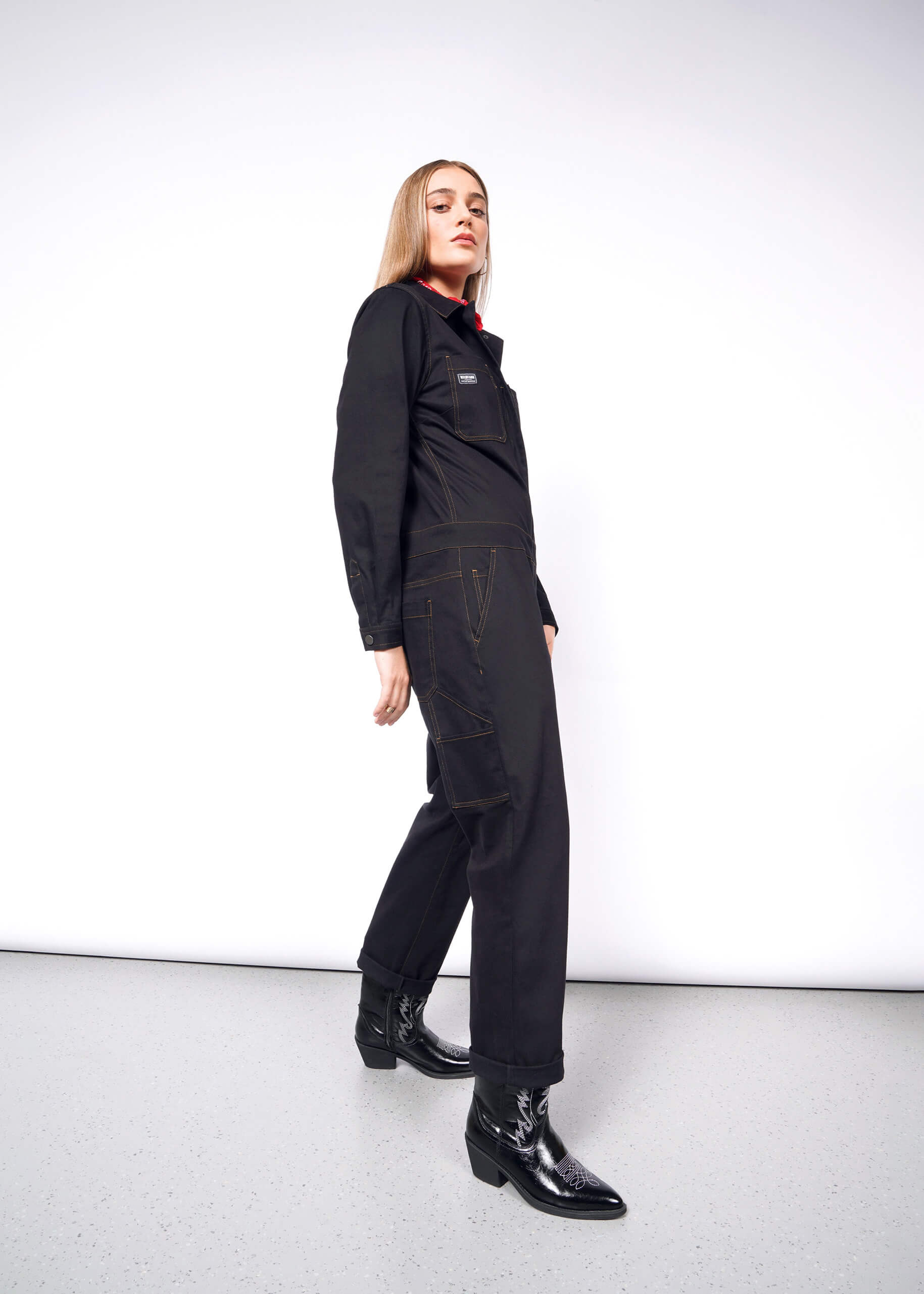 Against a white wall, the person exudes confidence in The Essential Long Sleeve Coverall and shiny boots. Long hair frames their face as they gaze into the camera, embodying a bold style.