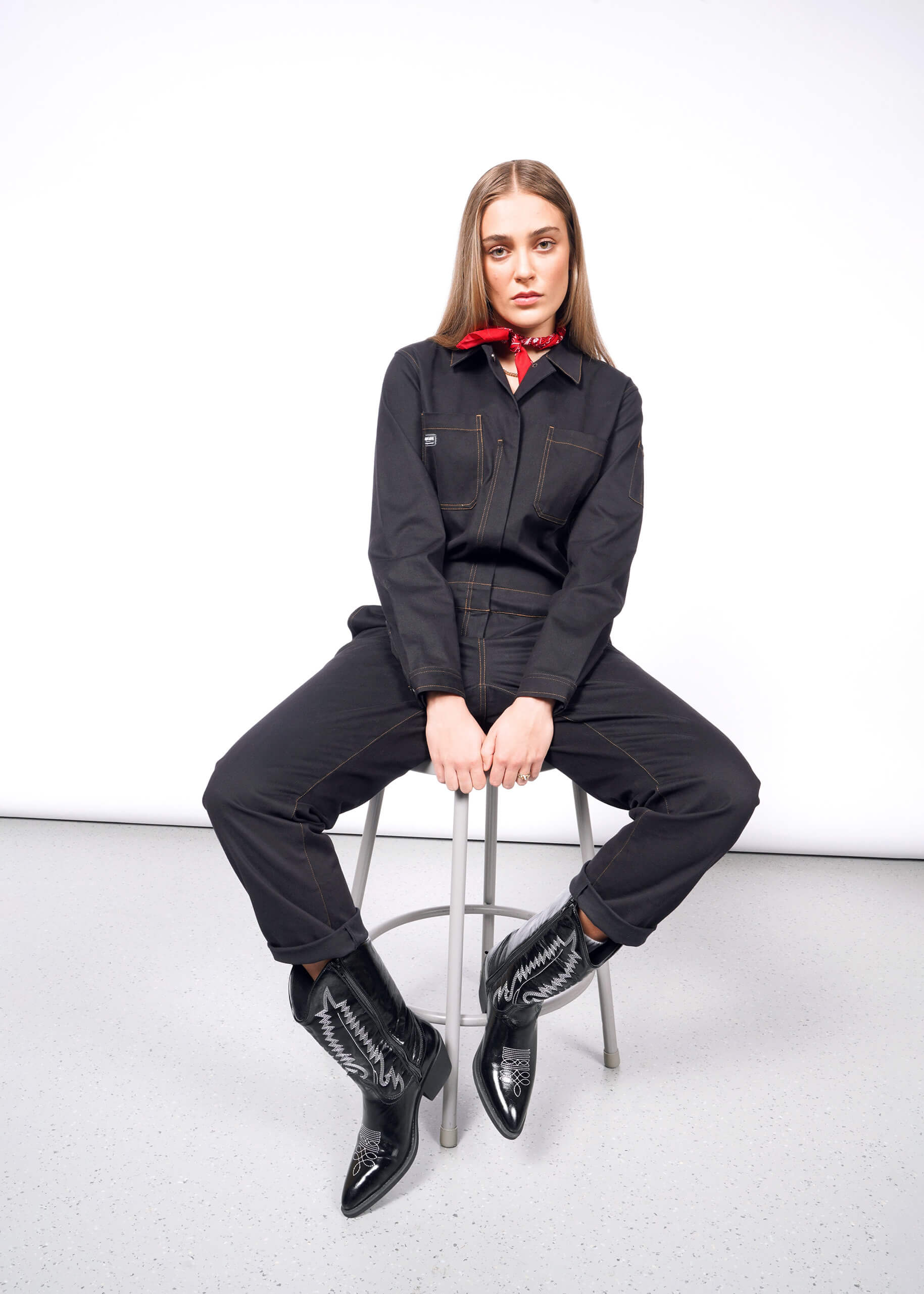 A person with long hair sits on a stool against a white background, wearing The Essential Long Sleeve Coverall in an oversized fit. Its crafted from brushed stretch cotton with visible stitching and pairs perfectly with their red bandana and black pointed cowboy boots, hands relaxed on their knees.