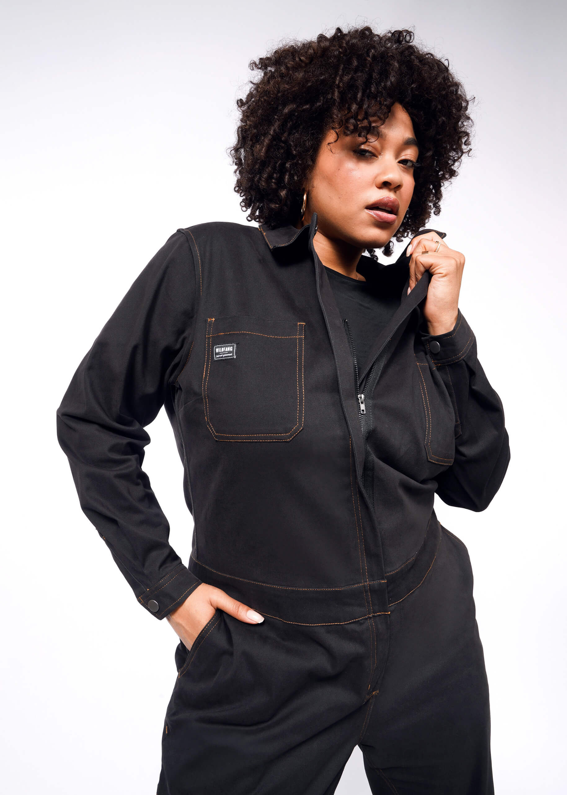 In a plain white setting, an individual with curly hair poses confidently in The Essential Long Sleeve Coverall. Made from brushed stretch cotton, this black coverall has brown stitching, an oversized fit, pockets for hands, and a small stitched label on the front.