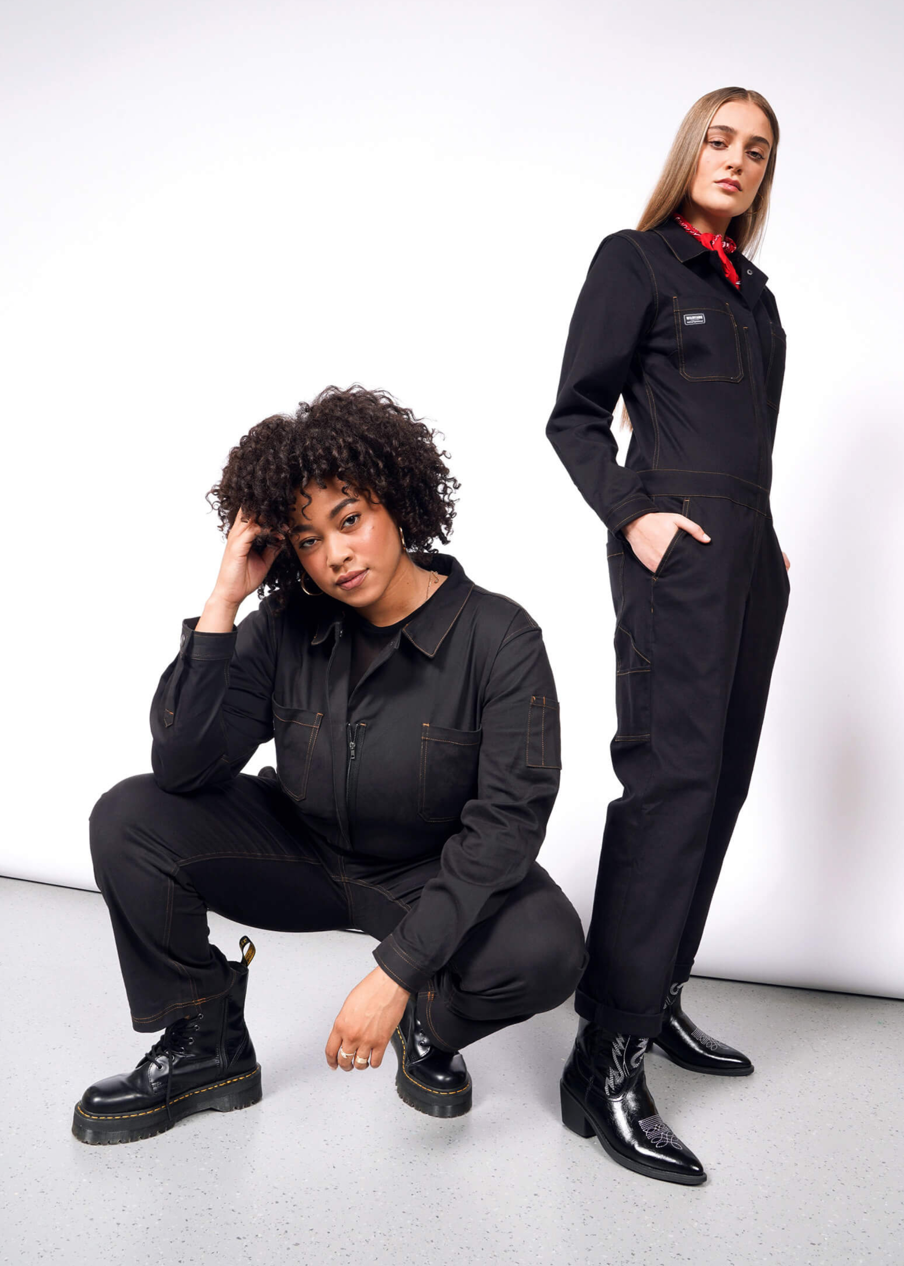 Two women wearing The Essential Long Sleeve Coverall in black pose against a white backdrop. One crouches with curly hair, while the other stands confidently with long, straight hair and a red bandana, both showcasing an oversized fit in brushed stretch cotton.