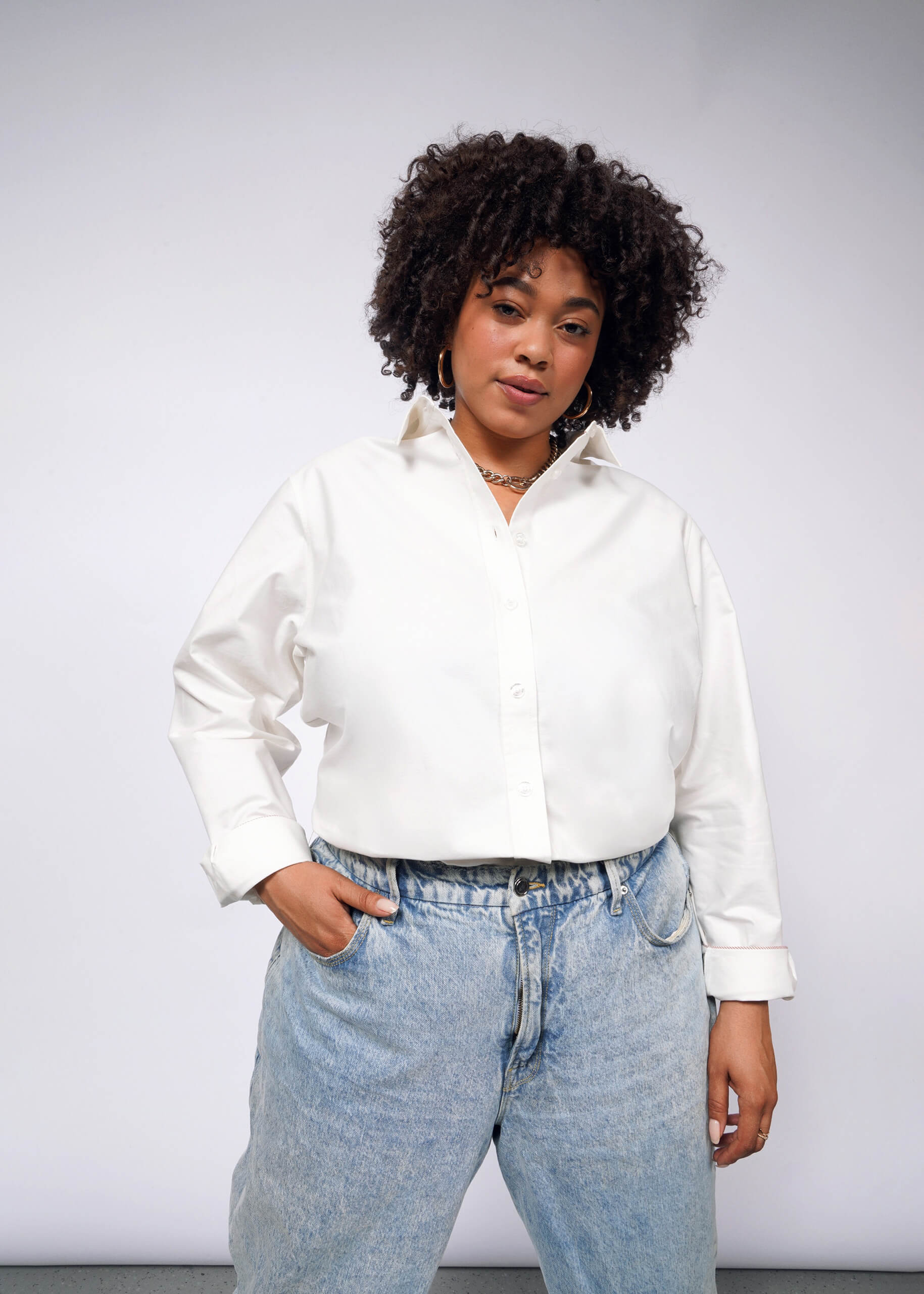 Against a plain background, a person with curly hair confidently models The Essential Long Sleeve Oxford Button Up, tailored for a perfect fit in 100% cotton, pairing it with light blue jeans with one hand casually in their pocket.