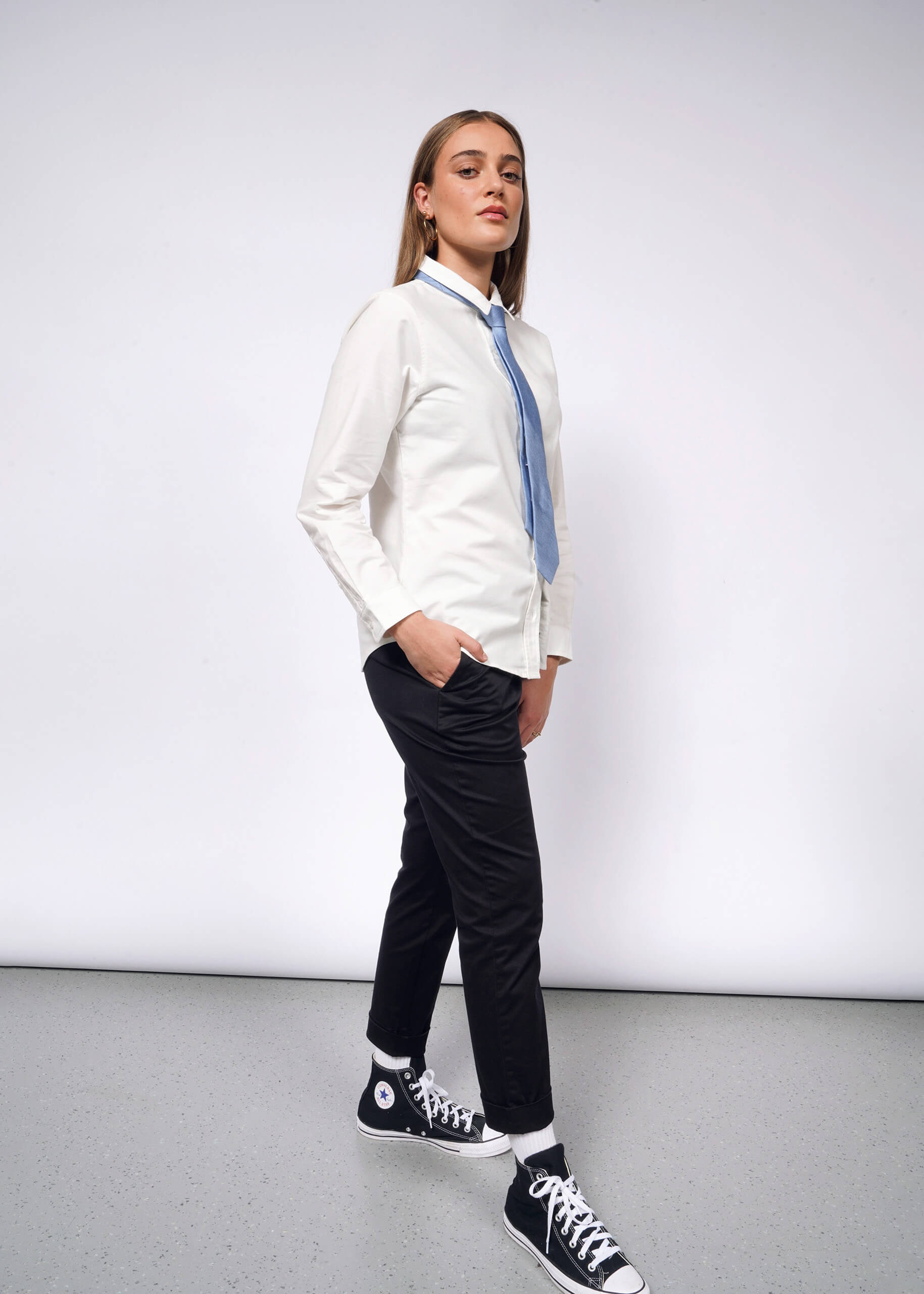 A person in The Essential Long Sleeve Oxford Button Up, a tailored white 100% cotton shirt, paired with a light blue tie, black pants, and high-top sneakers stands confidently against a plain backdrop. They have long hair and pose facing forward with a slight turn to the side.