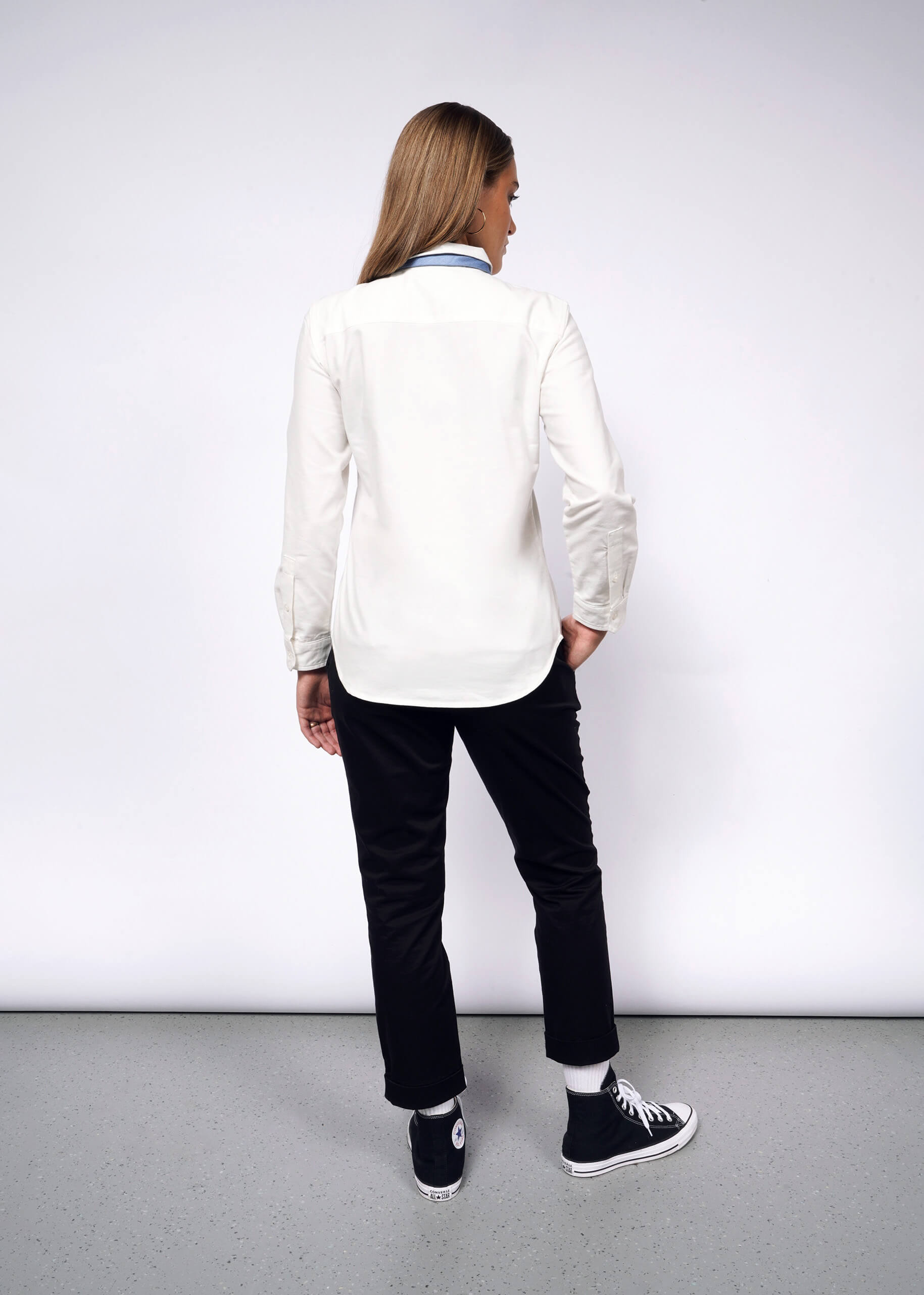 A person with long hair faces away from the camera, wearing The Essential Long Sleeve Oxford Button Up, crisp and tailored in white, along with black pants and high-top sneakers with white soles. The background is plain and light-colored.