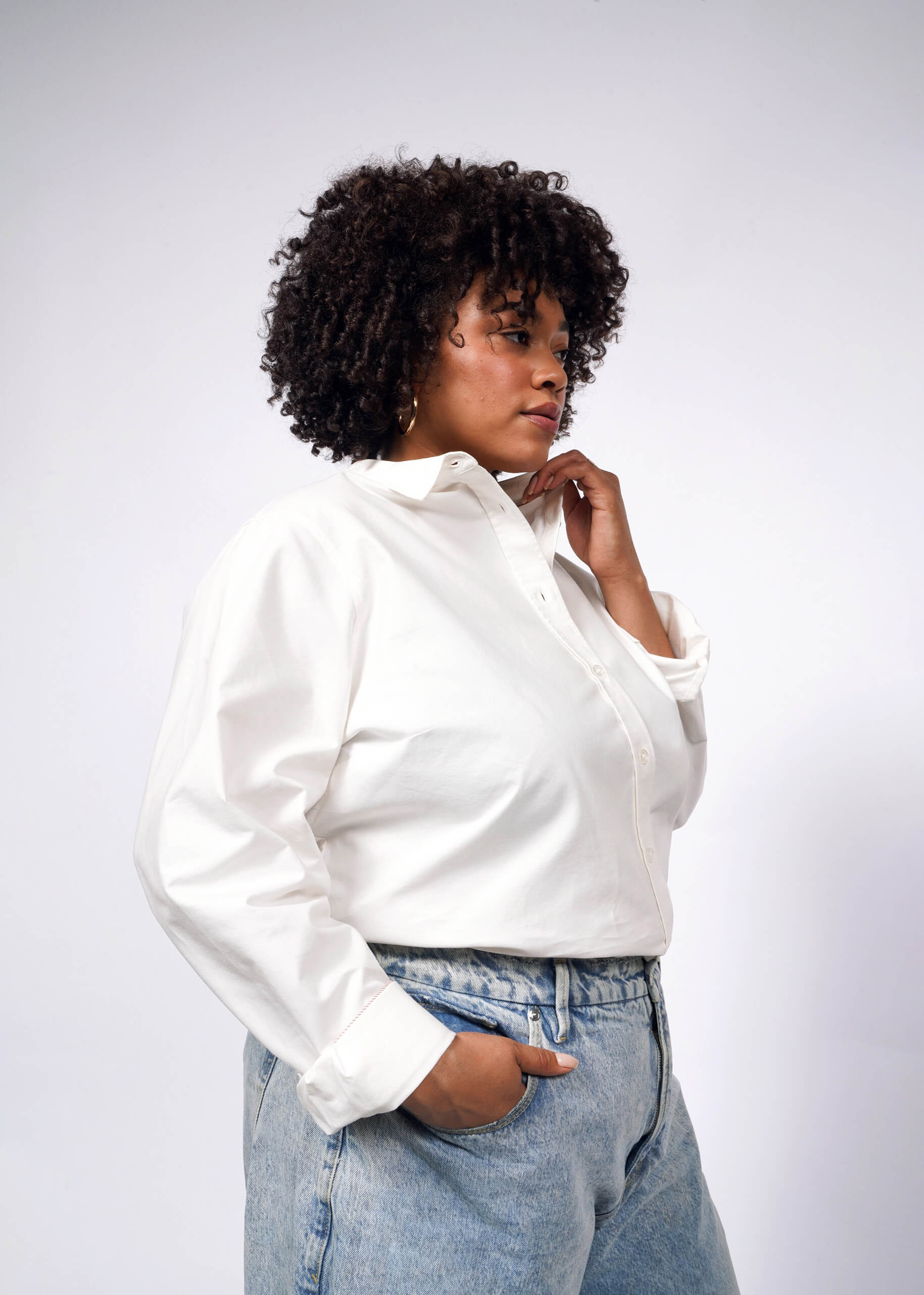 A person with curly hair stands against a plain background, wearing The Essential Long Sleeve Oxford Button Up shirt in tailored fit, paired with 100% cotton light blue jeans. One hand rests in their pocket while the other touches their chin, as they gaze thoughtfully to the side.