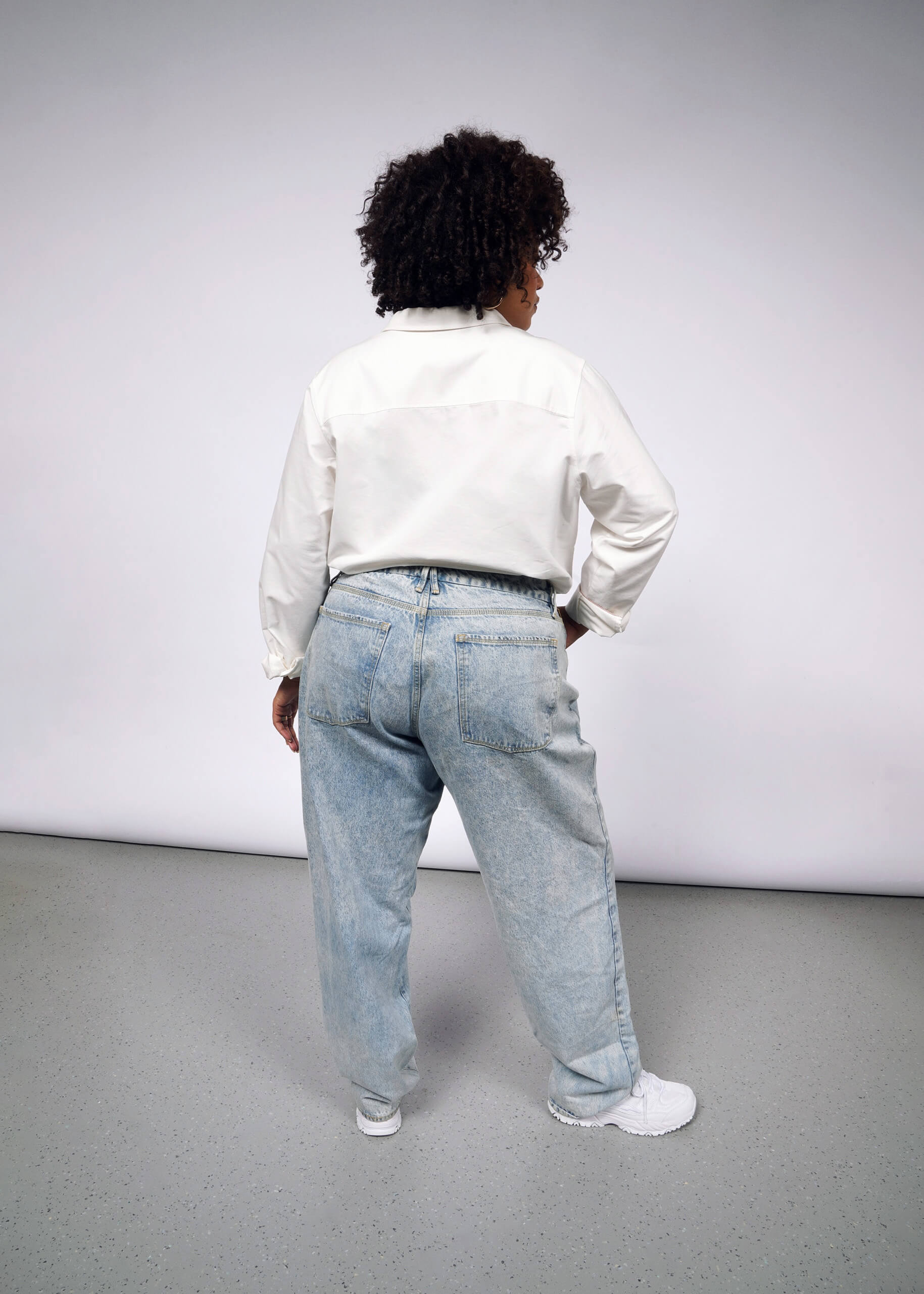 A person with curly hair stands with their back to the camera, wearing The Essential Long Sleeve Oxford Button Up in white, tailored fit, alongside light blue jeans and white sneakers. They pose casually against a plain background with one hand on their hip.