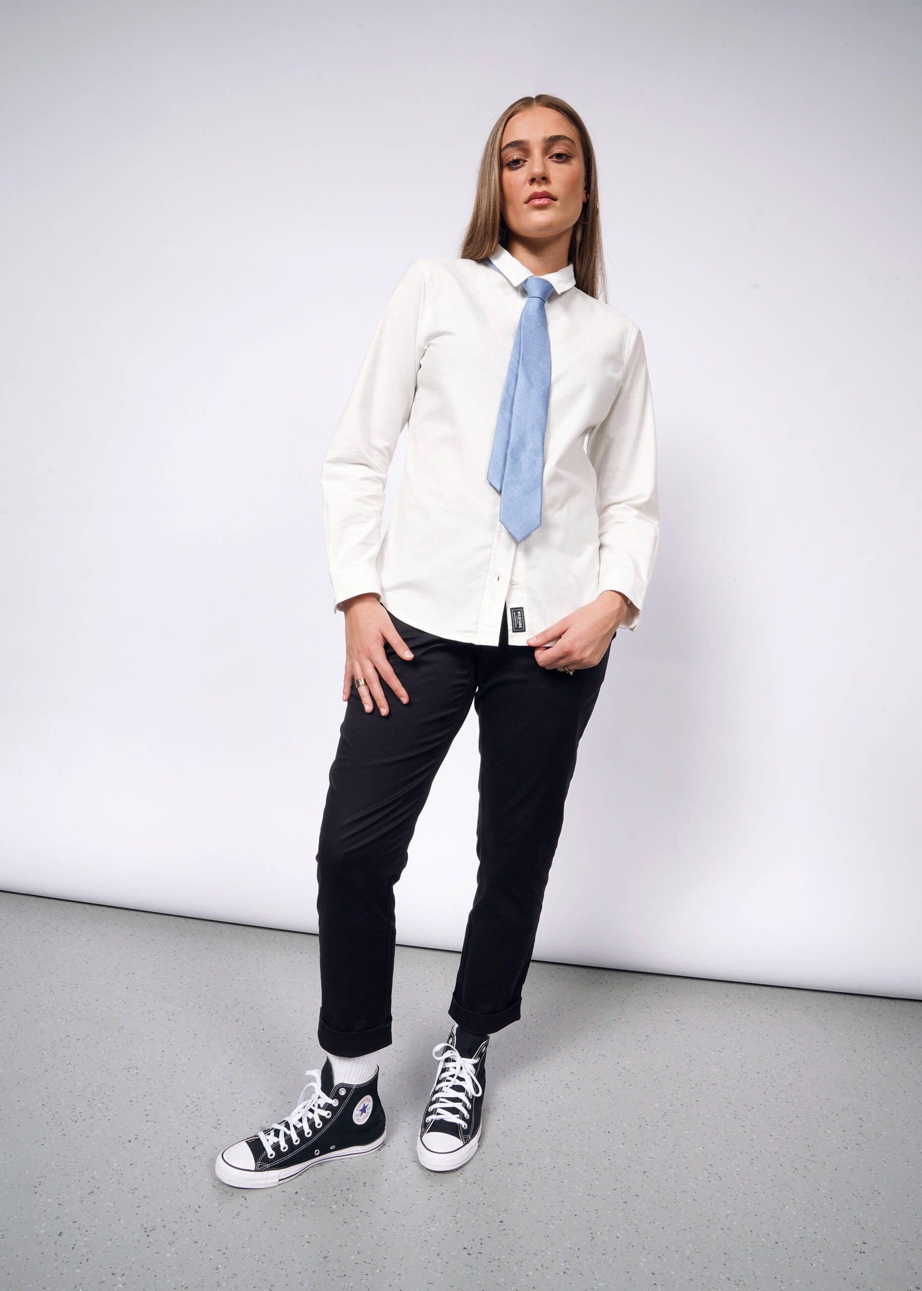 A person wearing The Essential Long Sleeve Oxford Button Up, paired with a blue tie, black pants, and black high-top sneakers, poses against a plain background. They stand with one foot slightly forward and their hands by their sides.