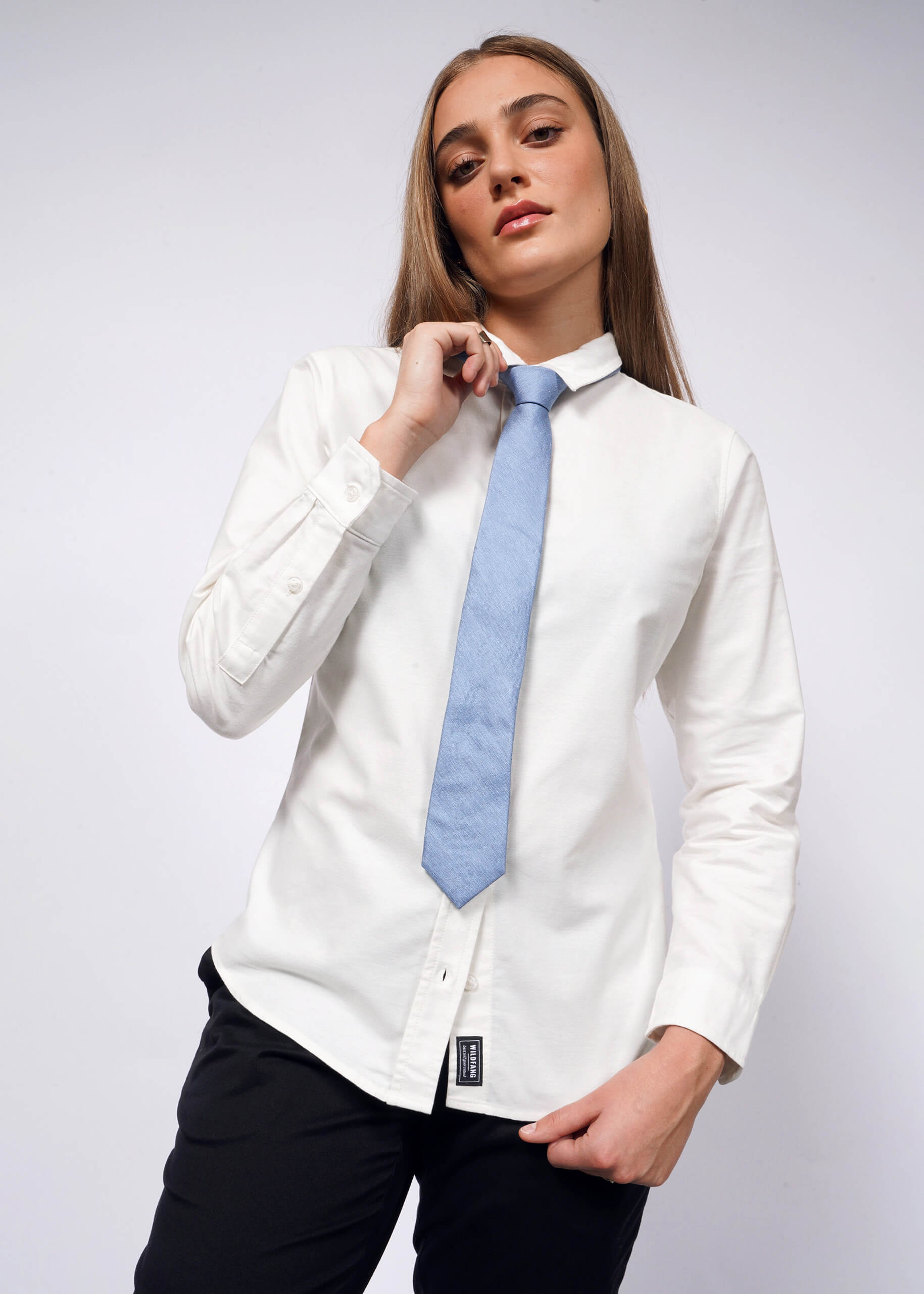 A person with long hair confidently stands against a plain background, wearing The Essential Long Sleeve Oxford Button Up made of 100% cotton, along with a light blue tie and black pants. One hand adjusts the tie.