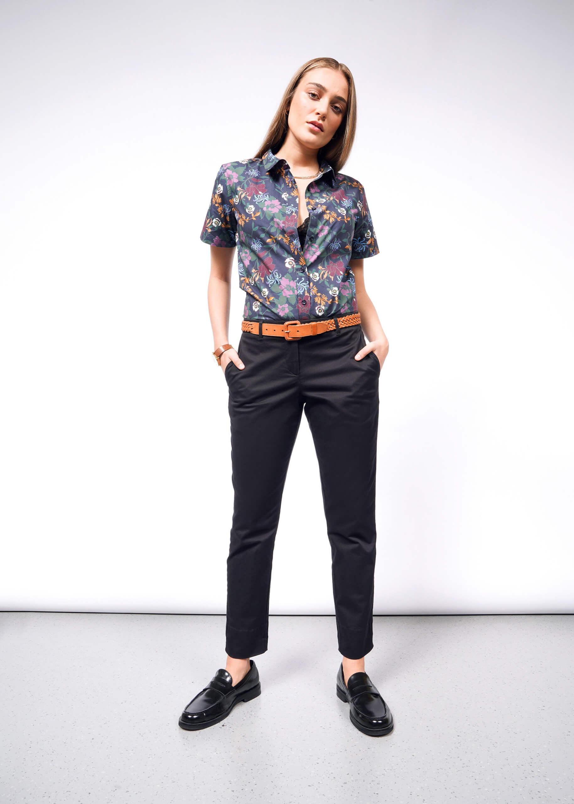 The woman stands confidently in a floral short-sleeve shirt and The Essential Trouser in relaxed mid-rise black. Black loafers and a brown belt with a bracelet complete the look. Her hands rest in her pockets against a plain light gray wall and floor, highlighting her poised stance.