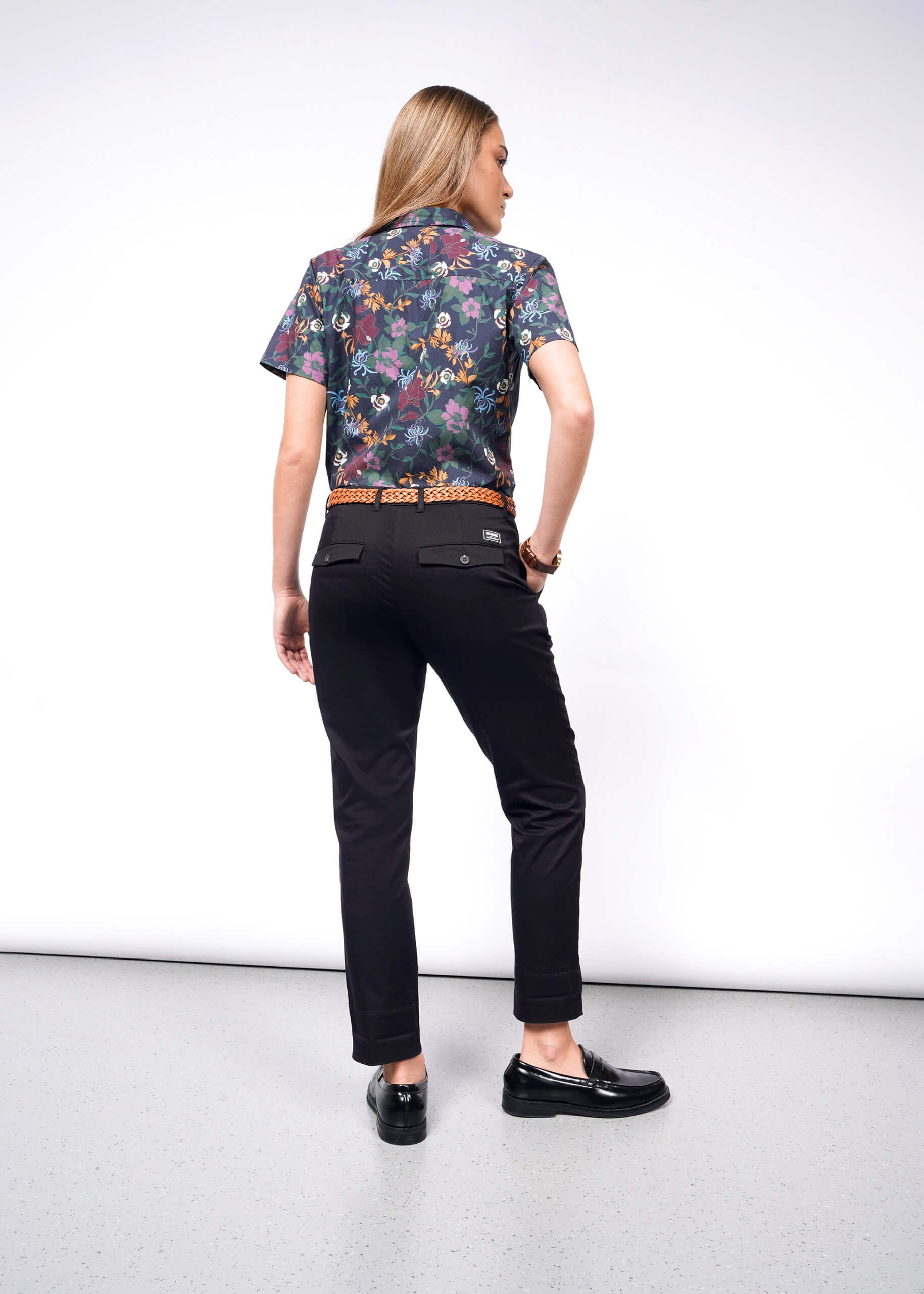 A person with long hair stands facing away, showcasing a floral short-sleeve shirt and The Essential Trouser in black with matching shoes. They stand on a light gray floor against a plain white background.