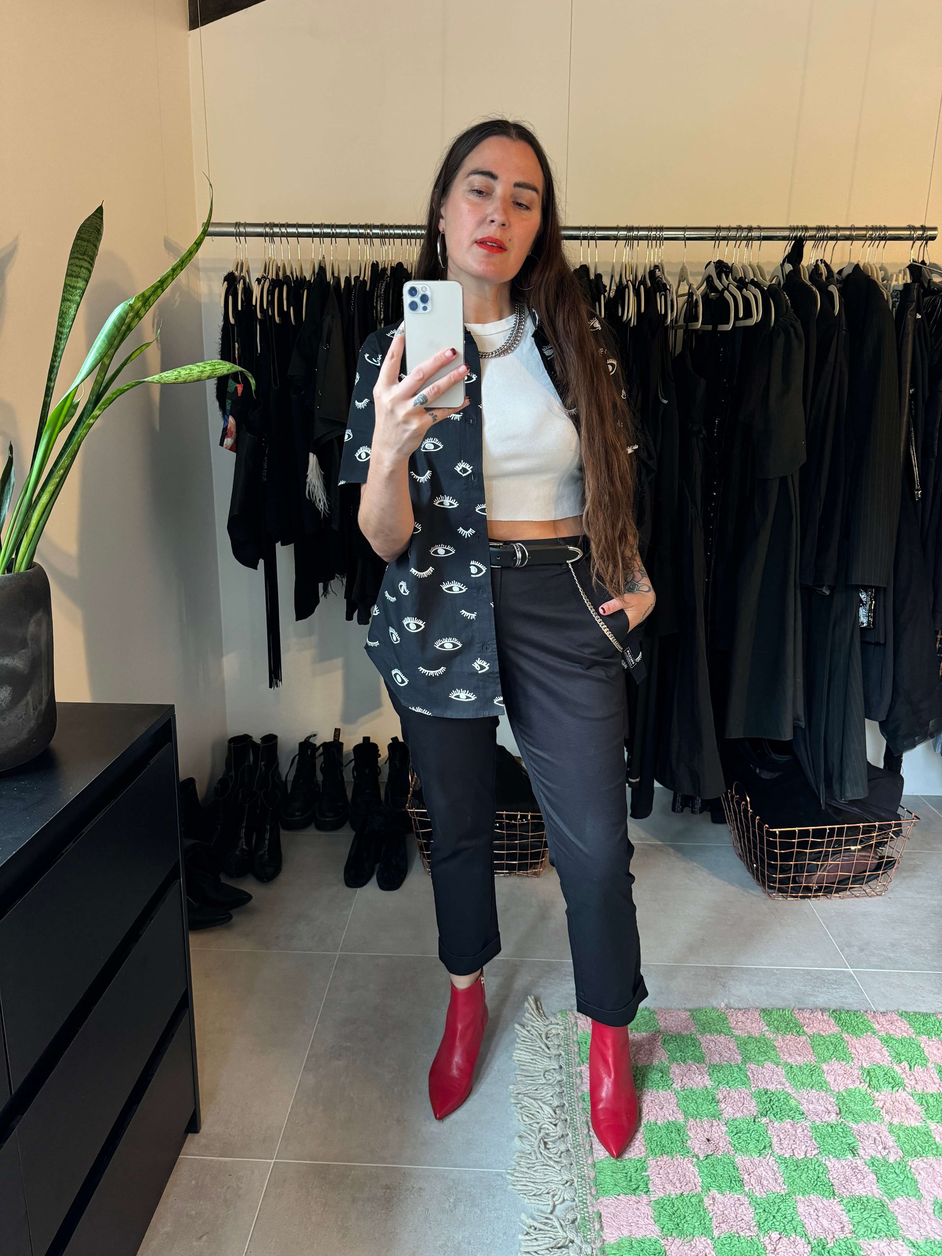 A woman takes a mirror selfie in a room with clothing racks. She wears The Essential Trouser in black and white paired with red boots, showcasing a relaxed mid-rise fit. Shes holding a phone, with a plant and rug completing the scene.