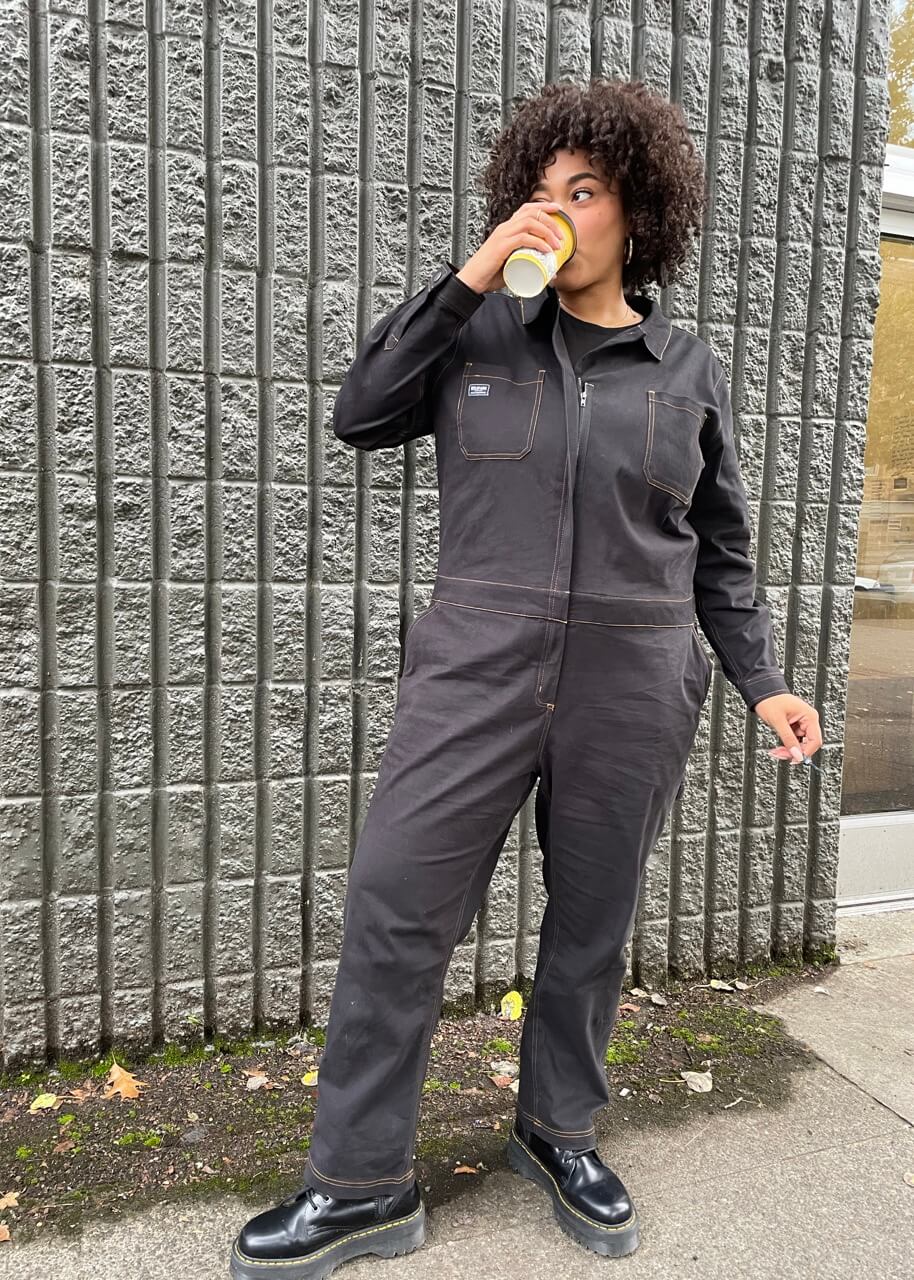 Wearing The Essential Long Sleeve Coverall in an oversized fit, a person with short curly hair sips from a cup, standing against a textured gray wall. They sport black shoes while scattered leaves on the ground enhance the cozy autumn vibe.