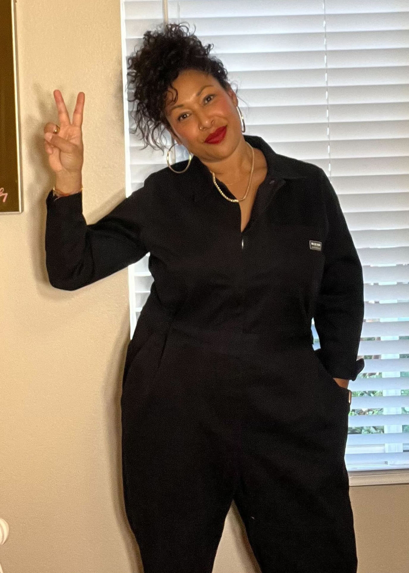 A woman wearing The Essential Long Sleeve High Waisted Coverall stands inside, smiling and making a peace sign with her right hand. Her curly hair is tied up, and shes accessorized with large hoop earrings and a gold necklace. A window with blinds is visible in the background.