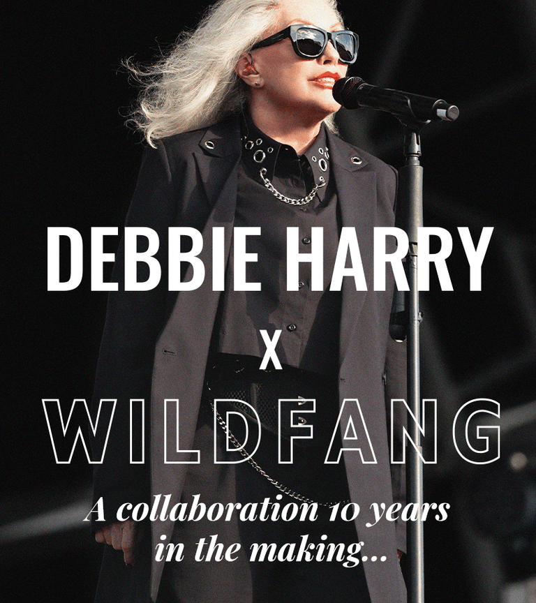 Close up of Debbie Harry with microphone stand on stage at her concert. She wears sunglasses and a suit and button up from the Debbie Harry x Wildfang collection. Text overlay reads DEBBIE HARRY X WILDFANG, a collaboration 10 years in the making...