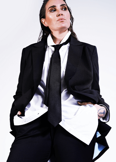 Person with long black hair wearing black suit, white button up, and black tie