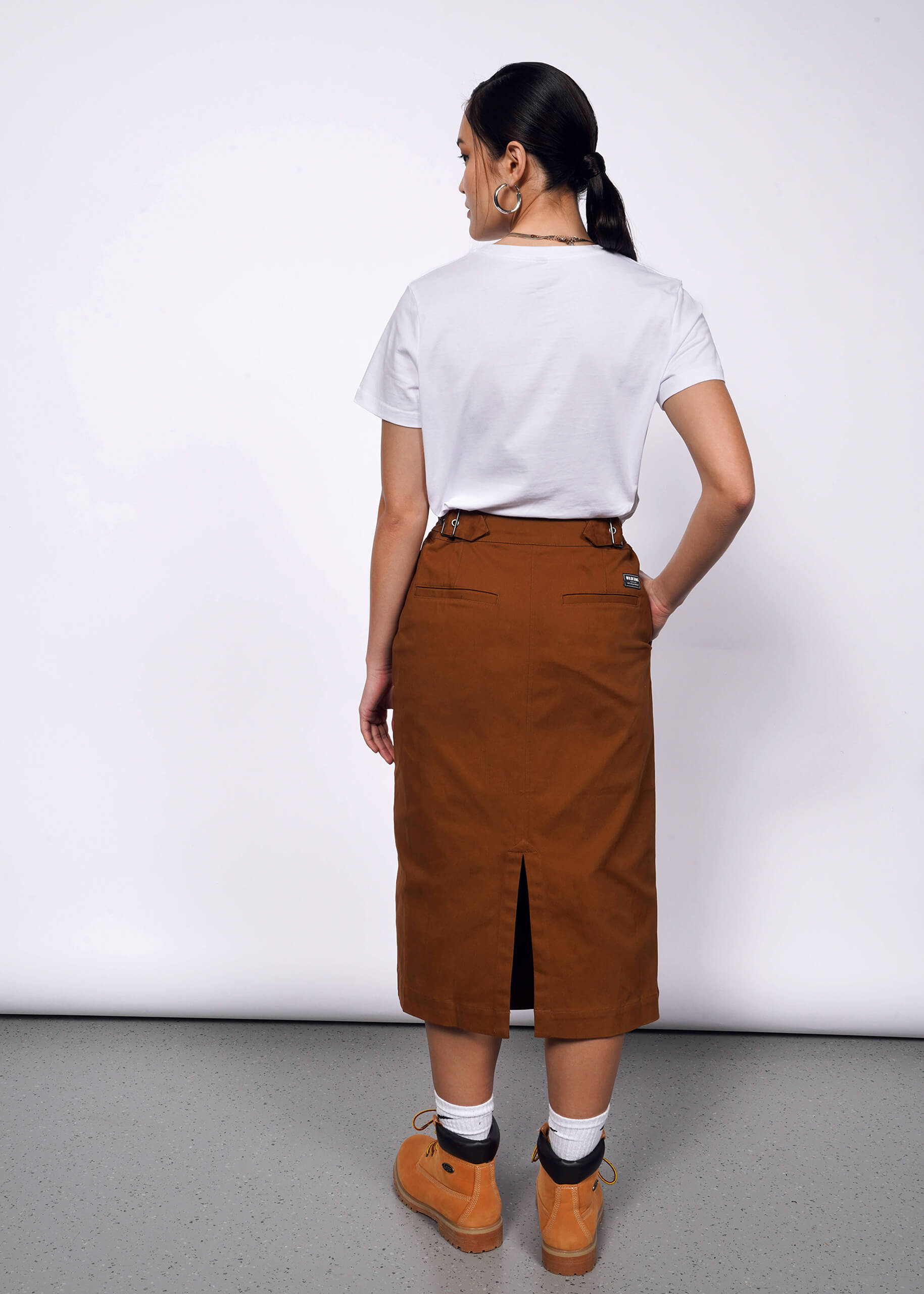 Back view of person with black hair tied back wearing white tshirt, brown skirt, and Timberland boots.