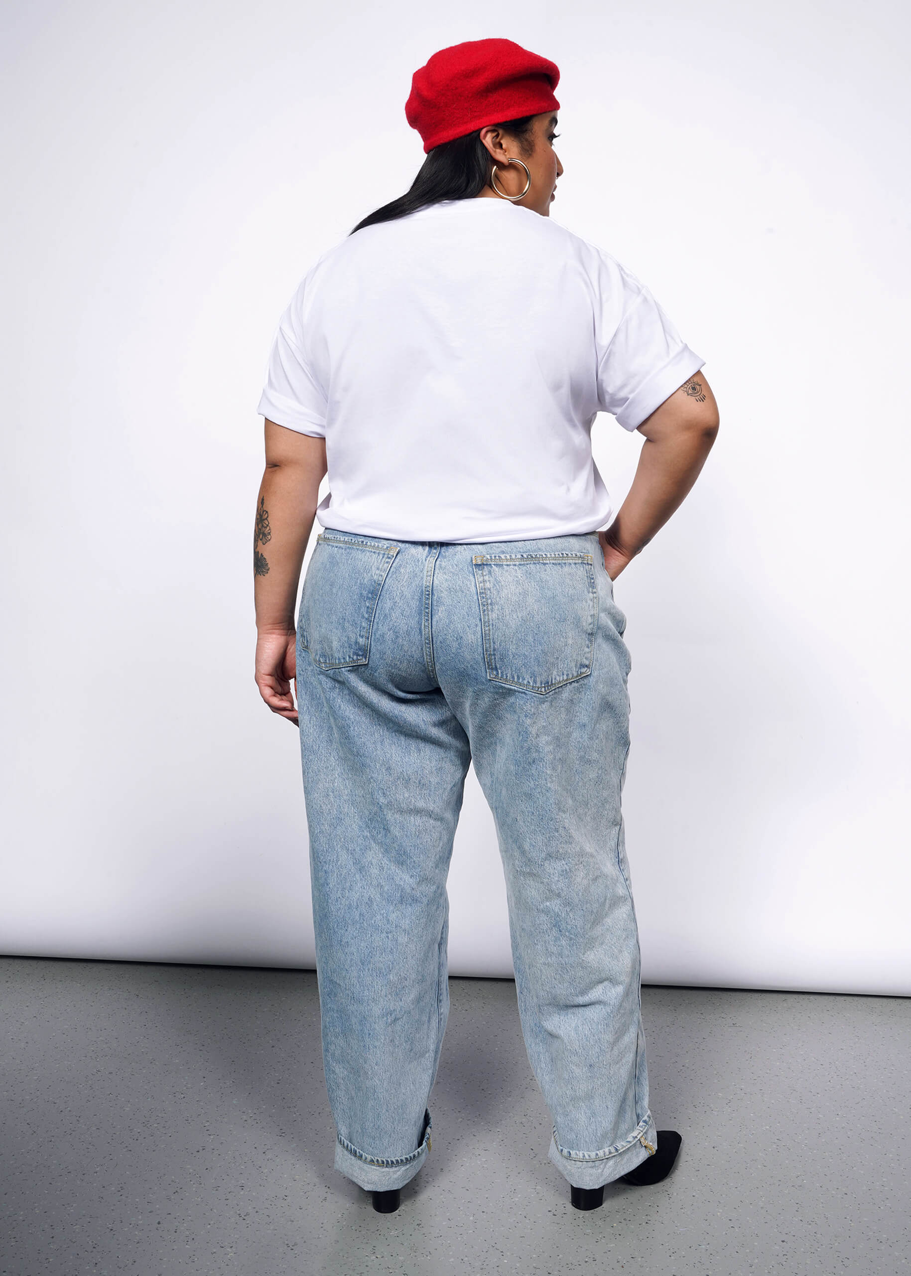 Back view of person with long black hair looking at camera wearing white tshirt, a red beret, jeans, and heeled boots.