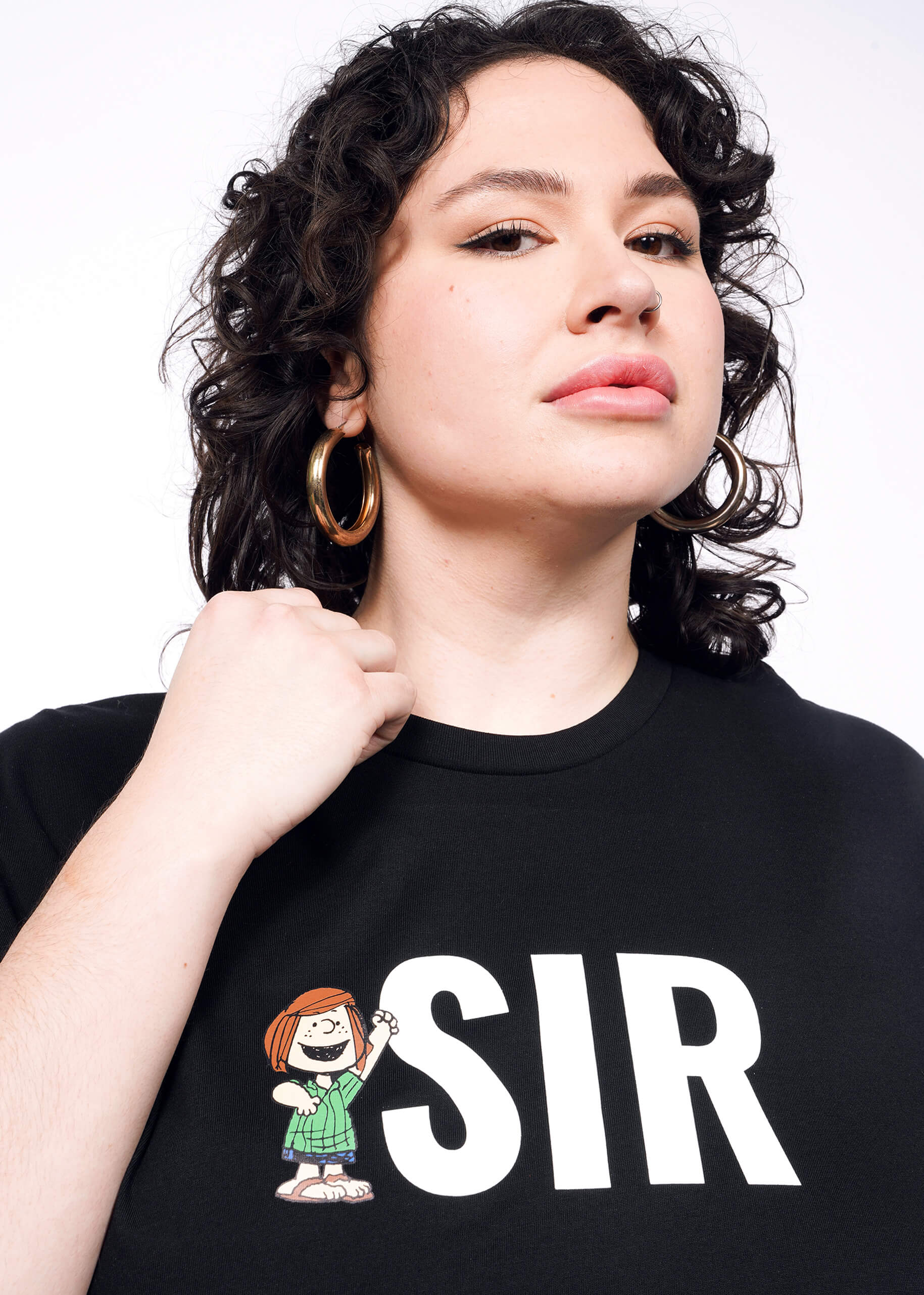 Close up of person with short curly black hair wearing hoop earrings and black tshirt with "SIR" and Peppermint Patty printed on it.