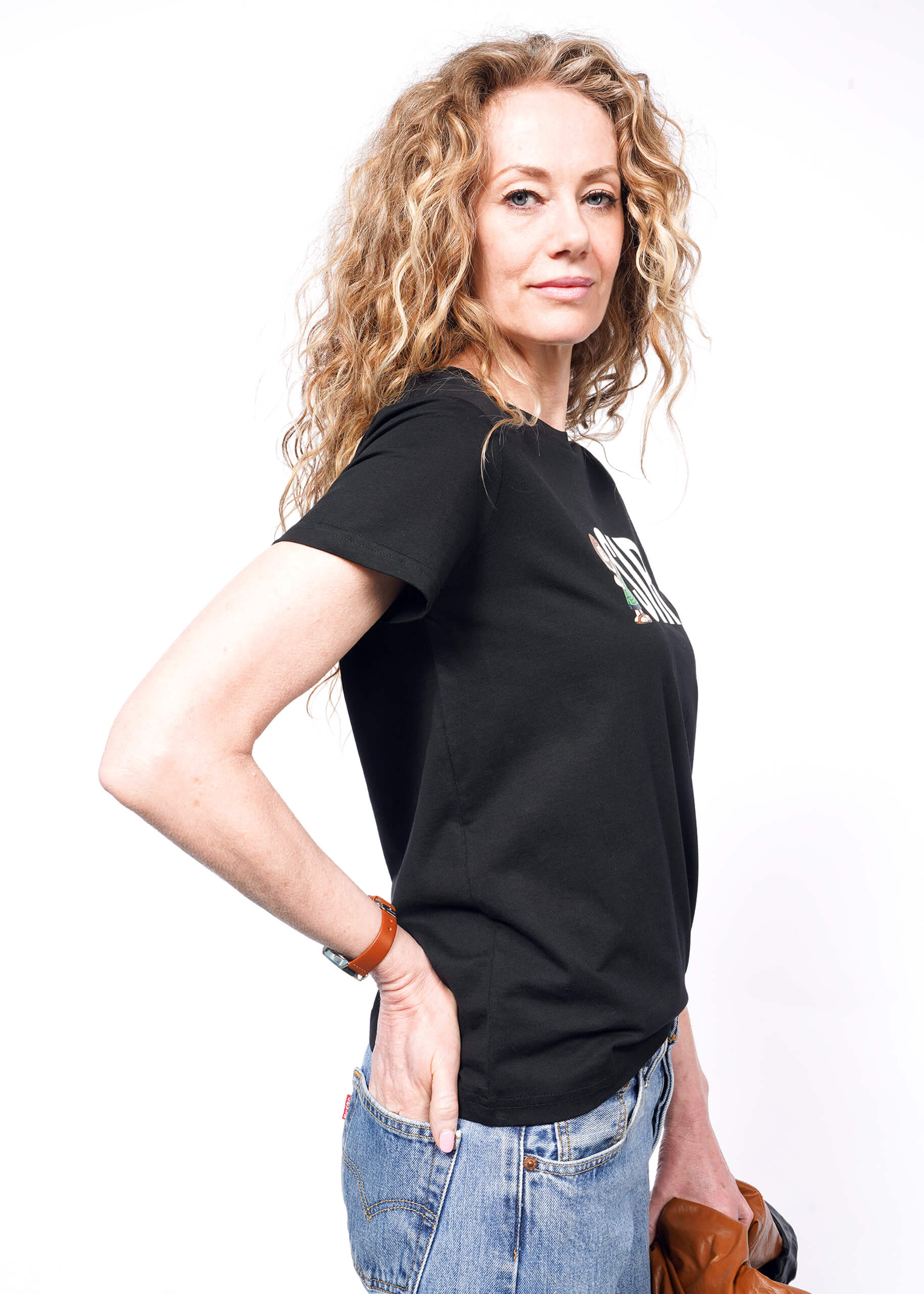 Side view of person with wavy blonde hair looking at camera while holding brown jacket wearing black tshirt with "SIR" and Peppermint Patty printed on it, and jeans.