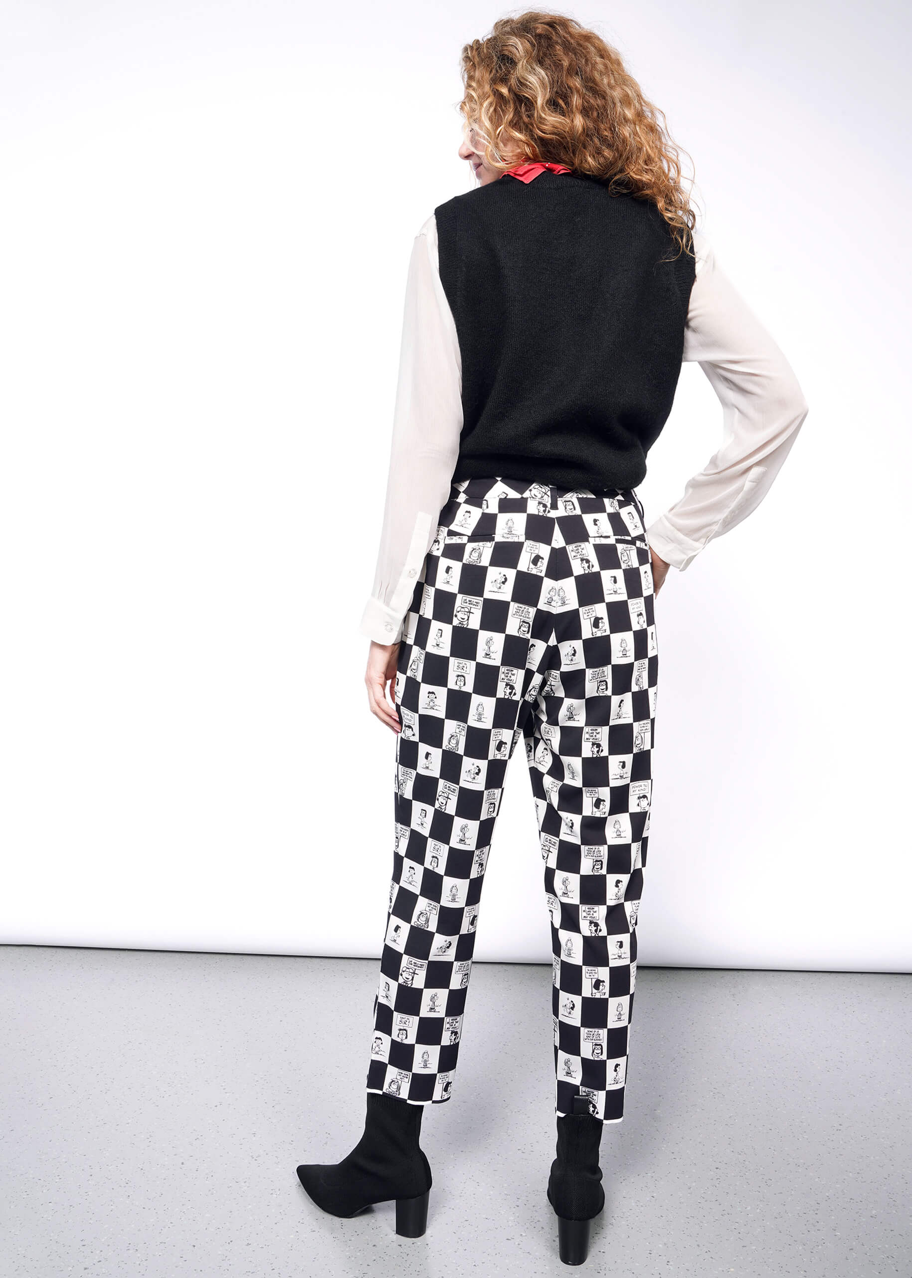 Back view of person with wavy blonde hair wearing white long sleeve button up under black sweater vest, a red bandana tied around their neck, black heeled boots, and the checkered black and white Slim Crop Pant from the Peanuts x Wildfang collection.