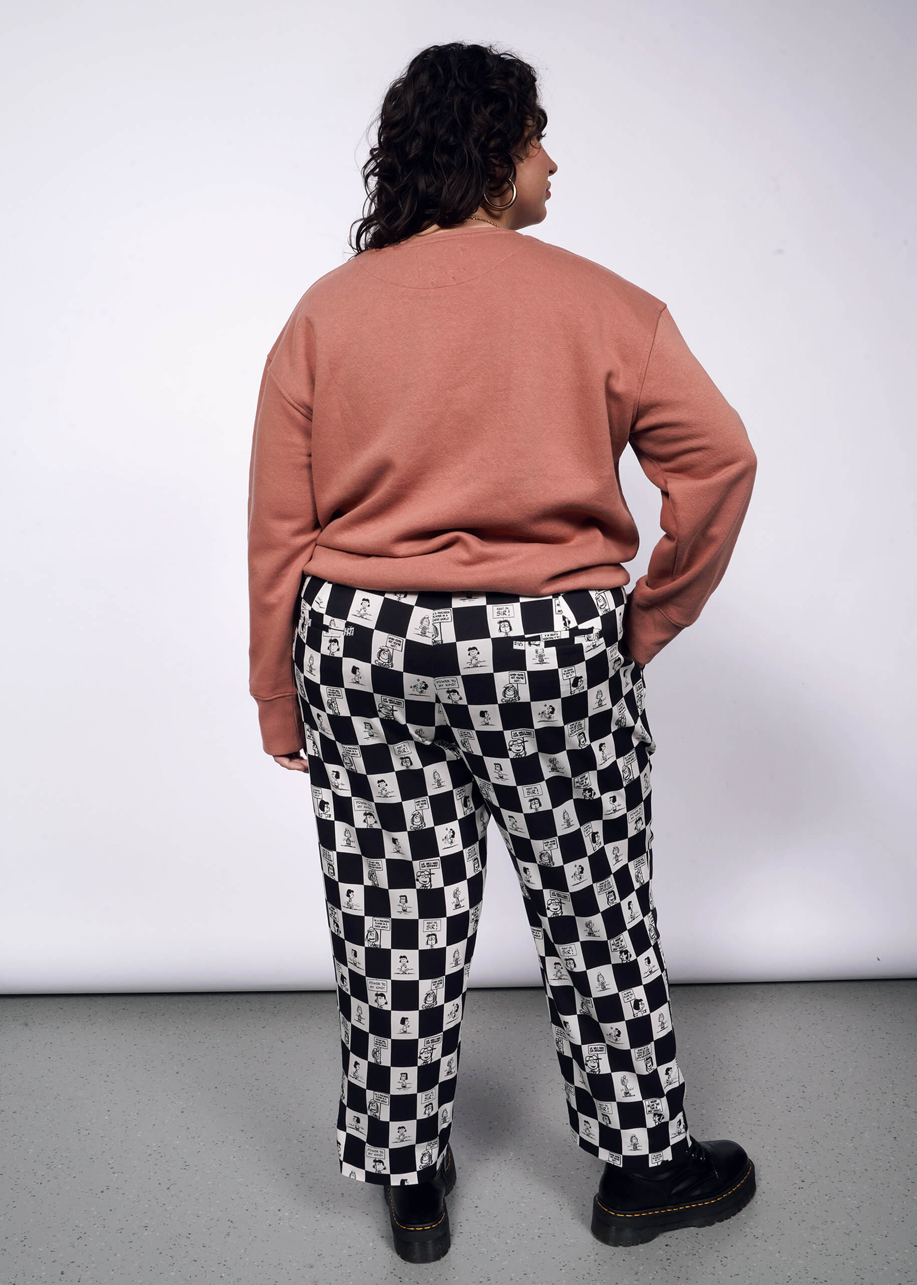 Back view of person with short curly black hair looking at camera with hand in pocket while wearing pink sweatshirt, black boots, and the checkered black and white Slim Crop Pant from the Peanuts x Wildfang collection.