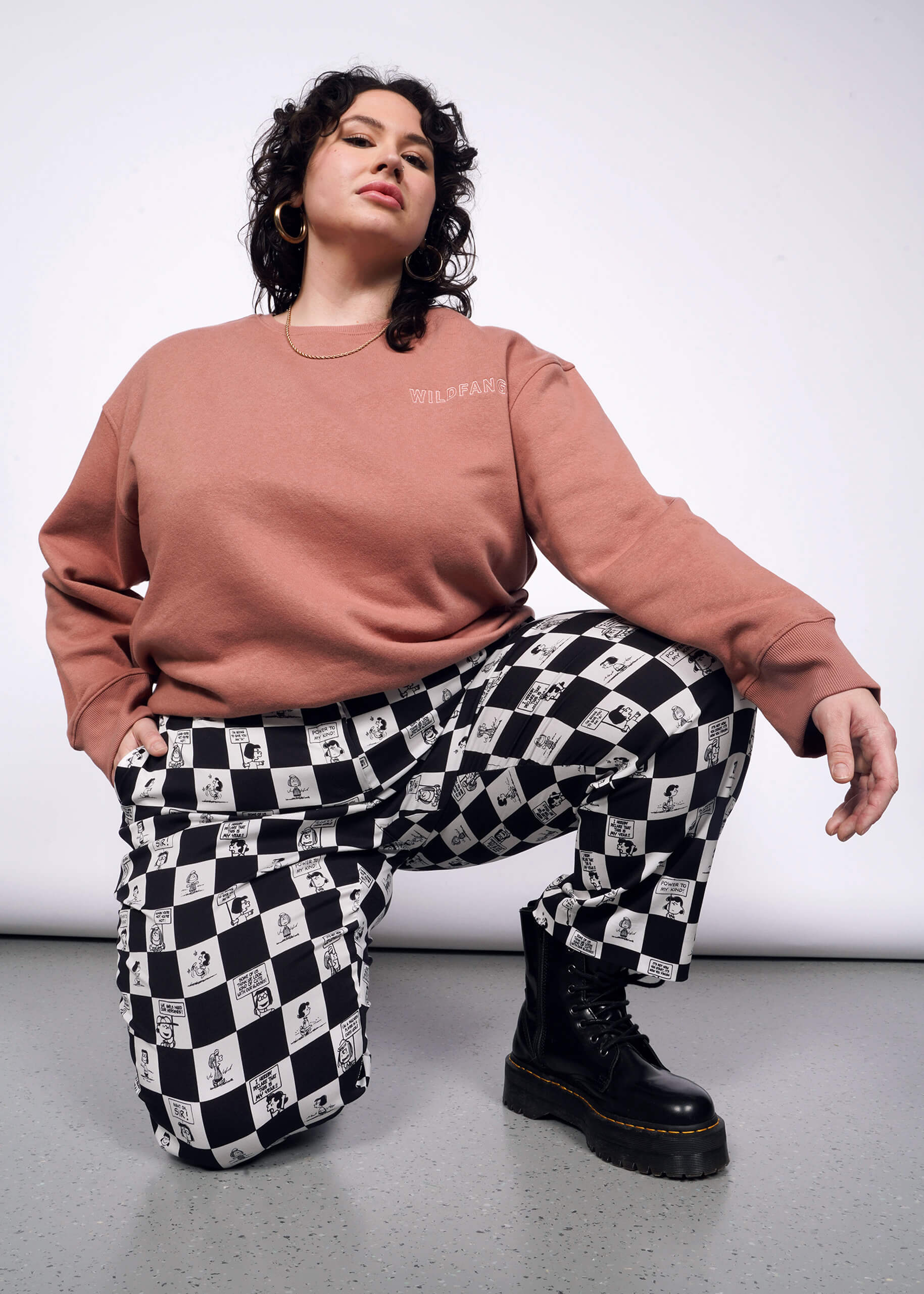 Person with short curly black hair bends down on one knee wearing pink sweatshirt, black boots, and the checkered black and white Slim Crop Pant from the Peanuts x Wildfang collection.