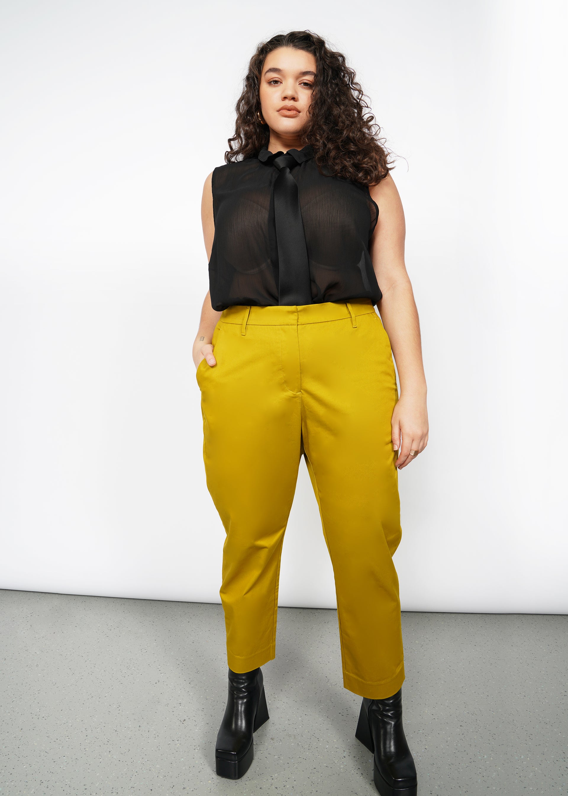 A woman with long curly hair wears a sheer black sleeveless blouse paired with The Essential Slim Crop Pant in mustard. She completes her look with black platform boots, standing confidently against a plain white background, her hands in her pockets.