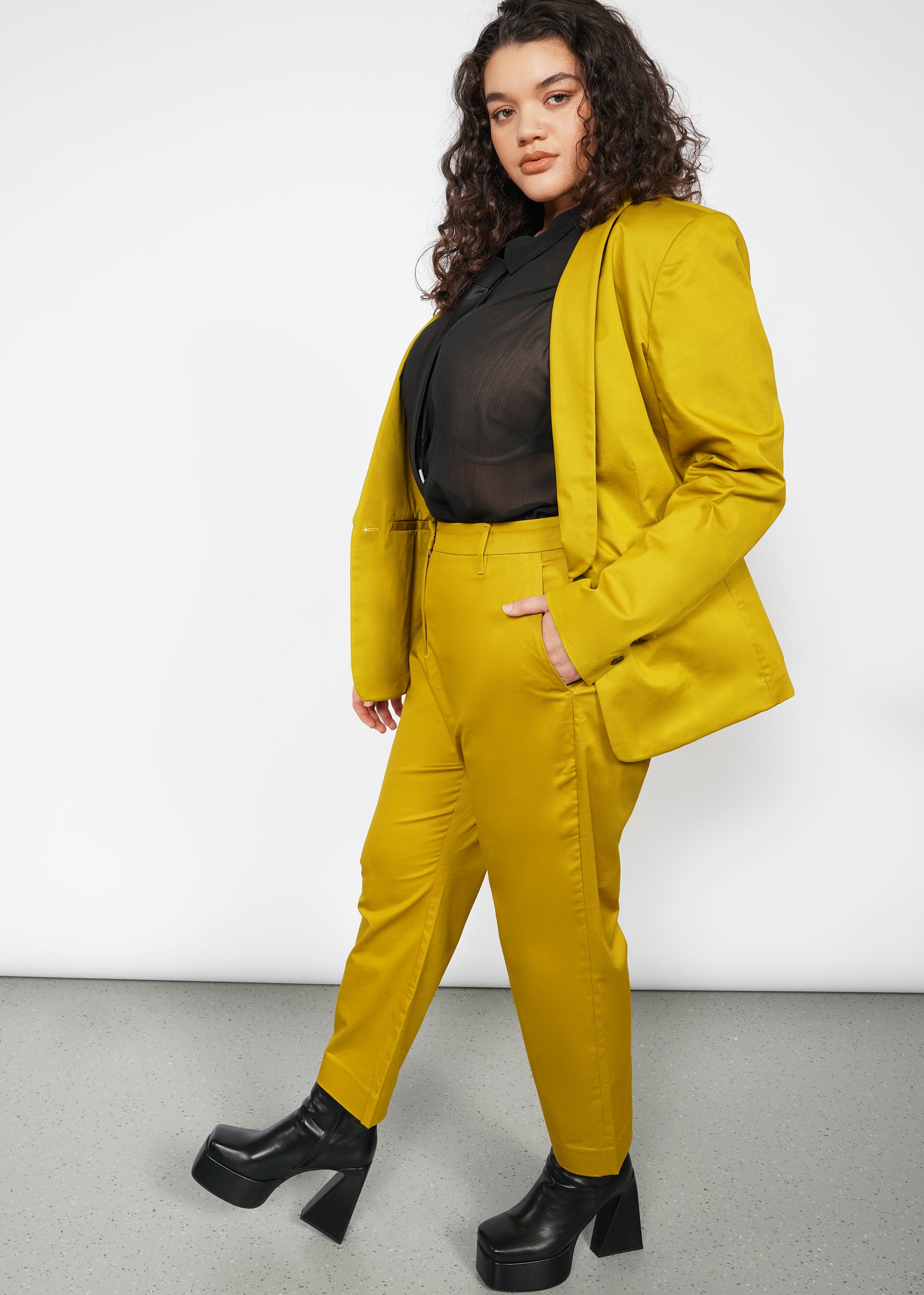 A person with long curly hair confidently wears a bright yellow suit featuring The Essential Slim Crop Pant, offering a breathable tailored mid-rise fit, paired with a sheer black blouse. They have black platform shoes and pose against a plain white background.