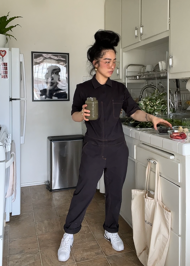 A person wearing The Essential Coverall in black and white sneakers stands in a kitchen, holding a jar of green juice. They lean against the countertop with a tote bag hanging from a drawer. A black-and-white photo and some plants decorate the background.