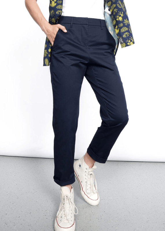 Dressed in The Essential Slim Crop Pant, navy blue and mid-rise, paired with white high-tops featuring star logos, the person stands confidently. A floral-patterned blazer adds flair as one hand rests casually in a pocket on the gray floor against a white background.